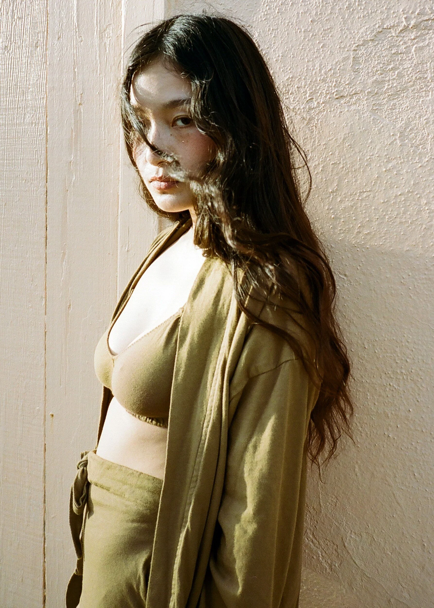 A model in an olive bra, open robe and drawstring trousers leans against a pale wall.