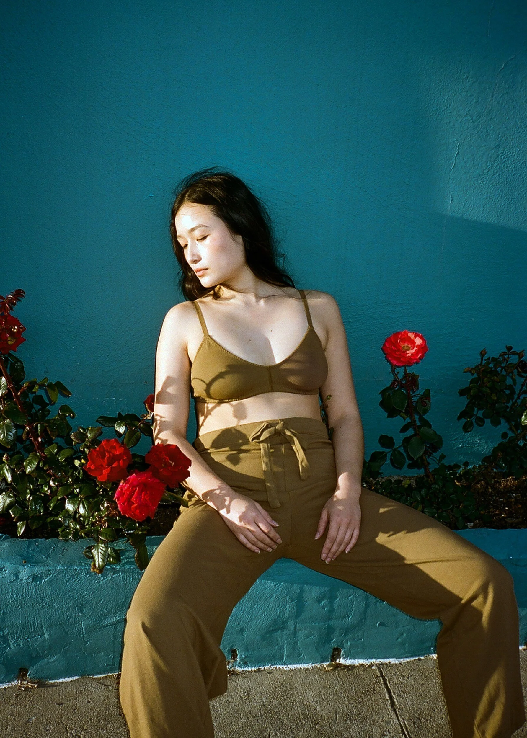 A model in a brown bra and drawstring trousers sits with legs apart among red roses against a blue wall.