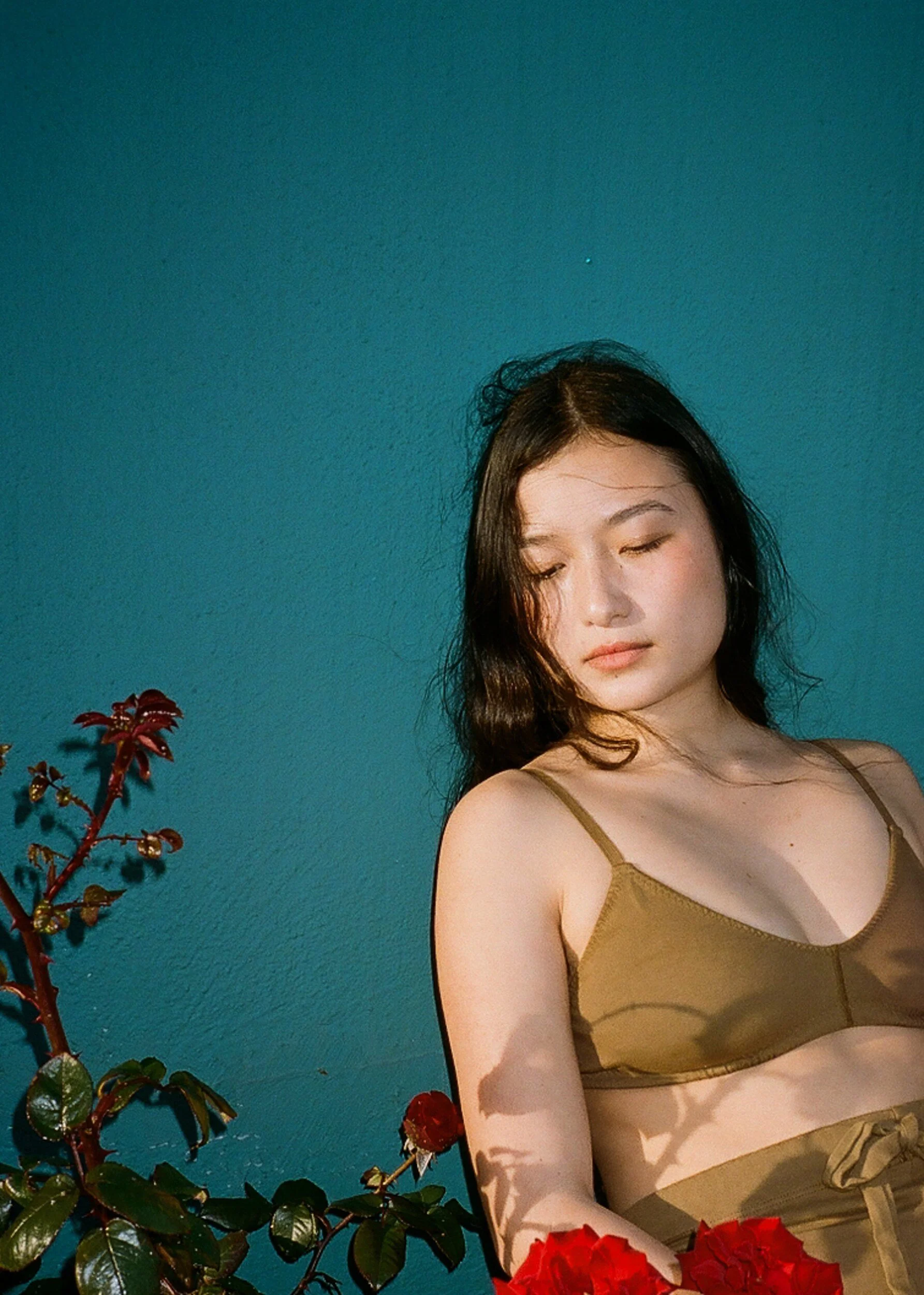 A model in a brown bra looks down beside red roses, with leaf shadows across their body.