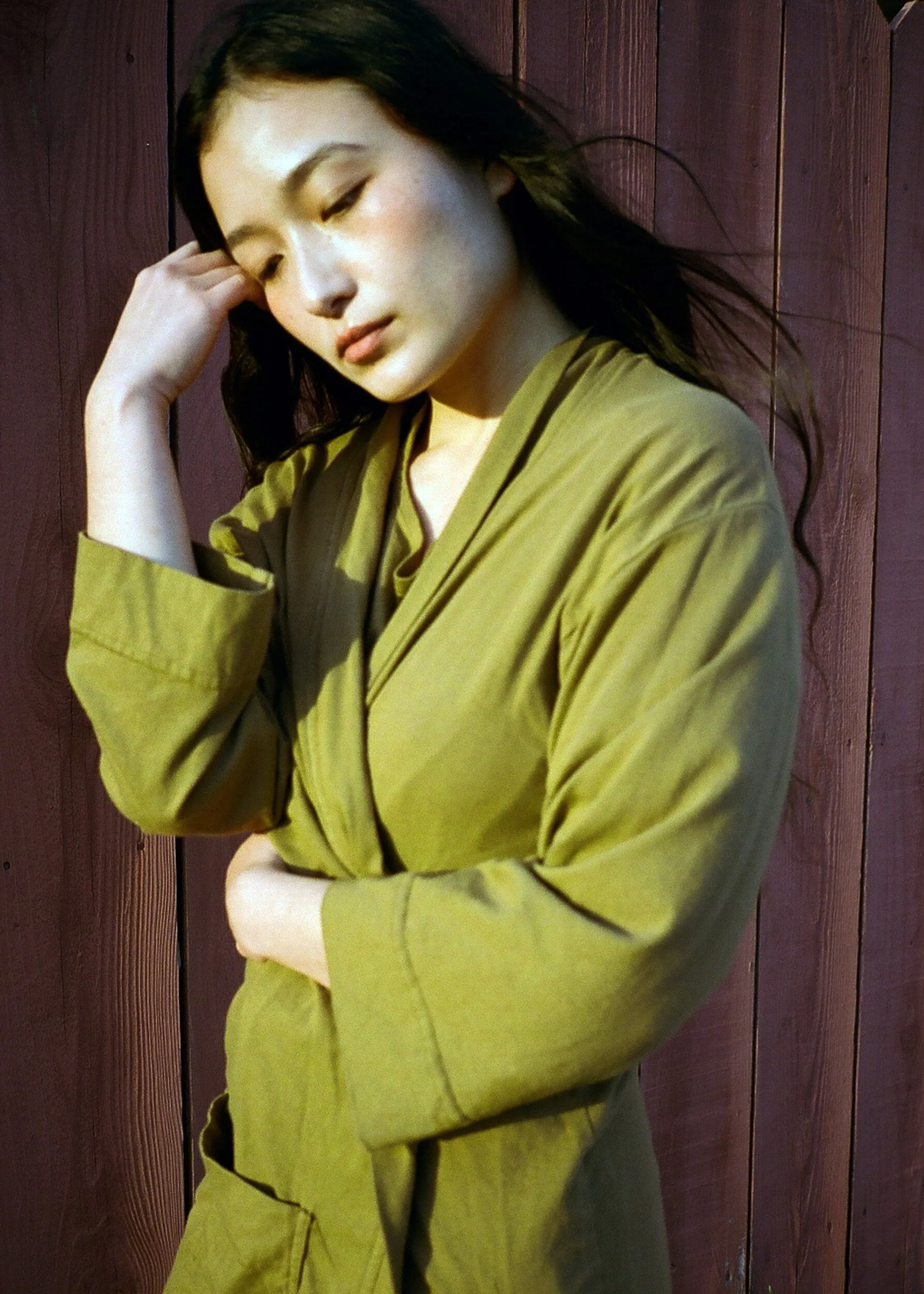 A model in an olive robe rests a hand at their temple beside a red wooden fence.