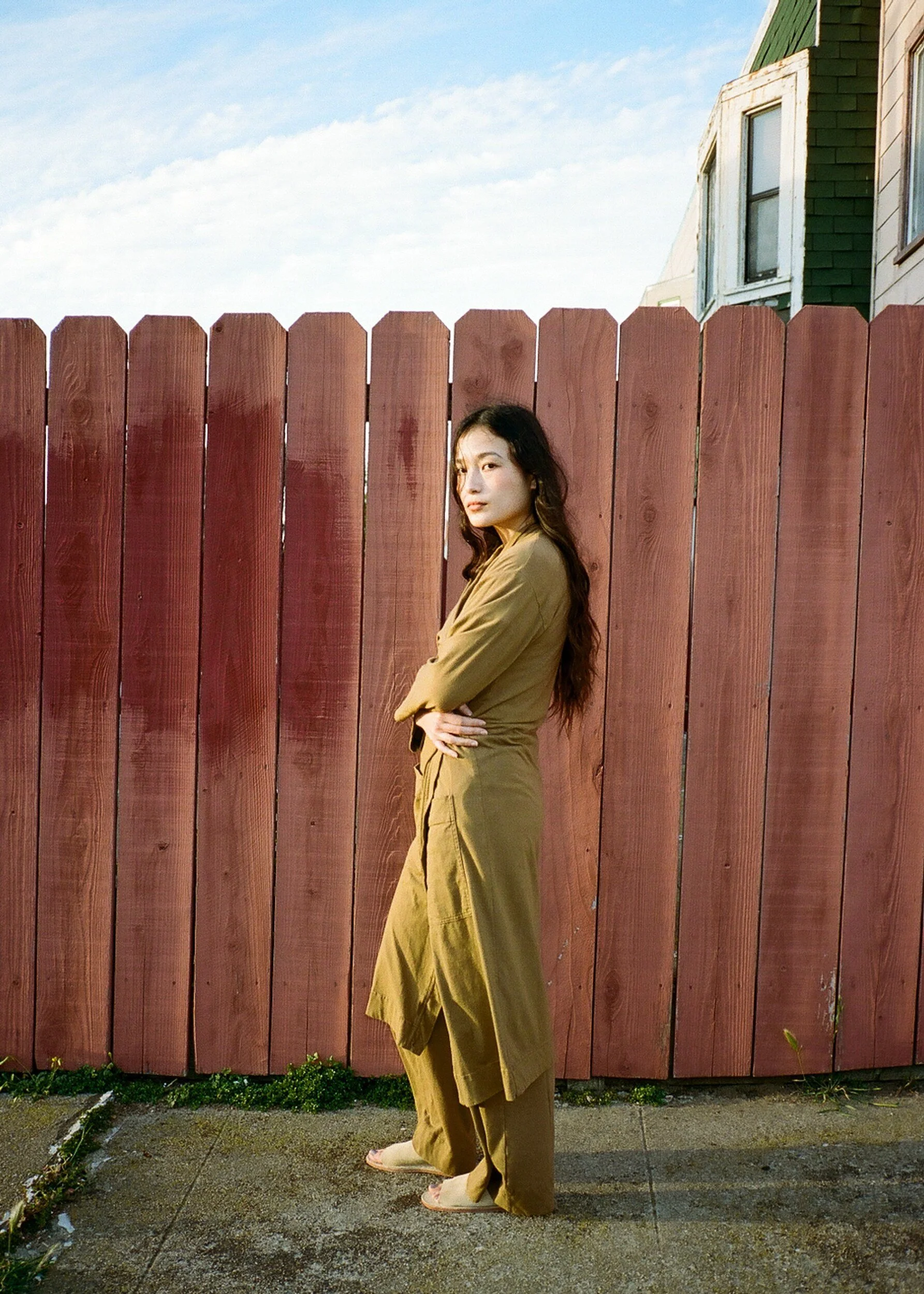 A barefoot model in an olive robe and trousers stands sideways beside a red wooden fence.