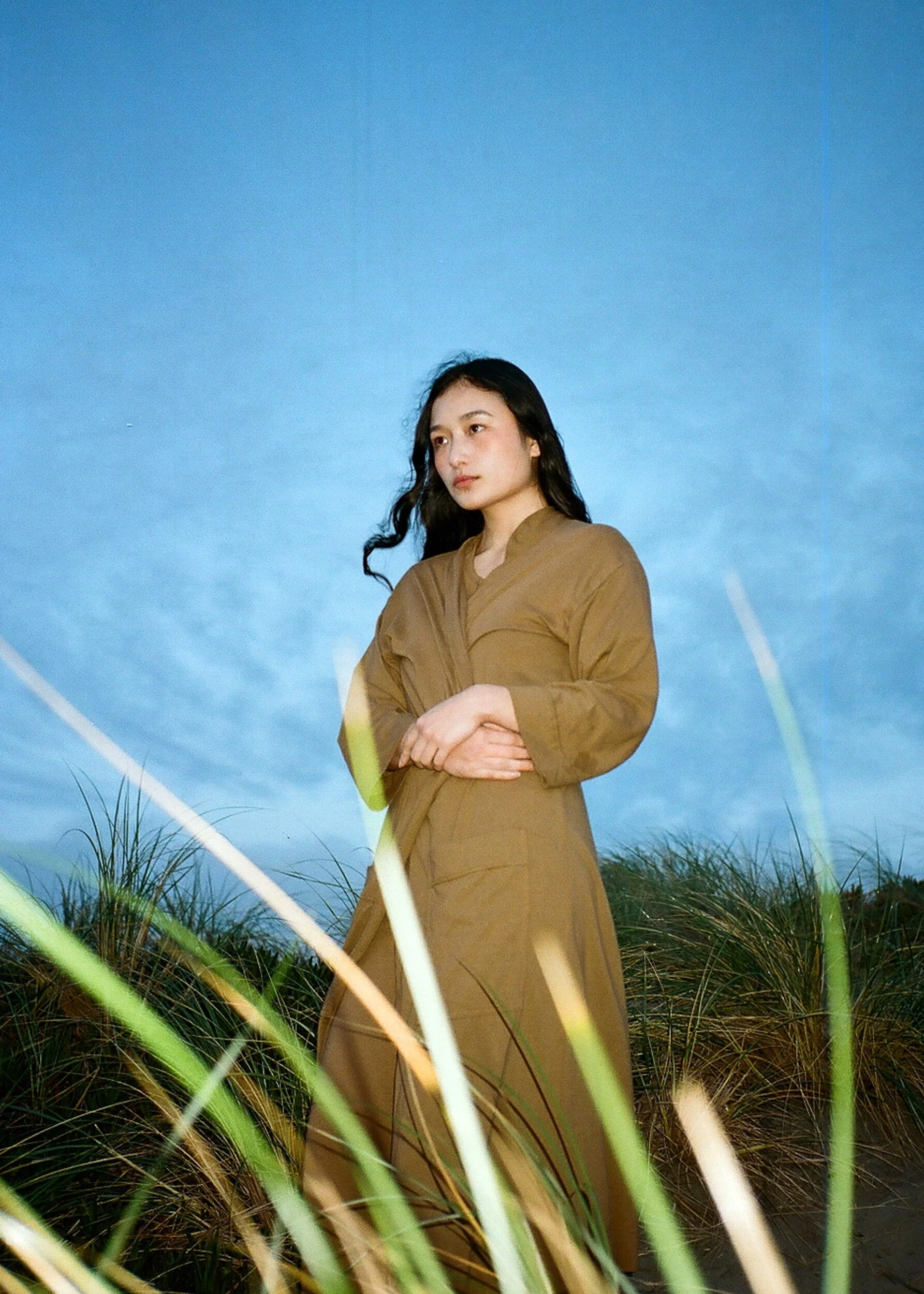 A model in a brown robe and trousers stands among tall grass against a bright blue backdrop.