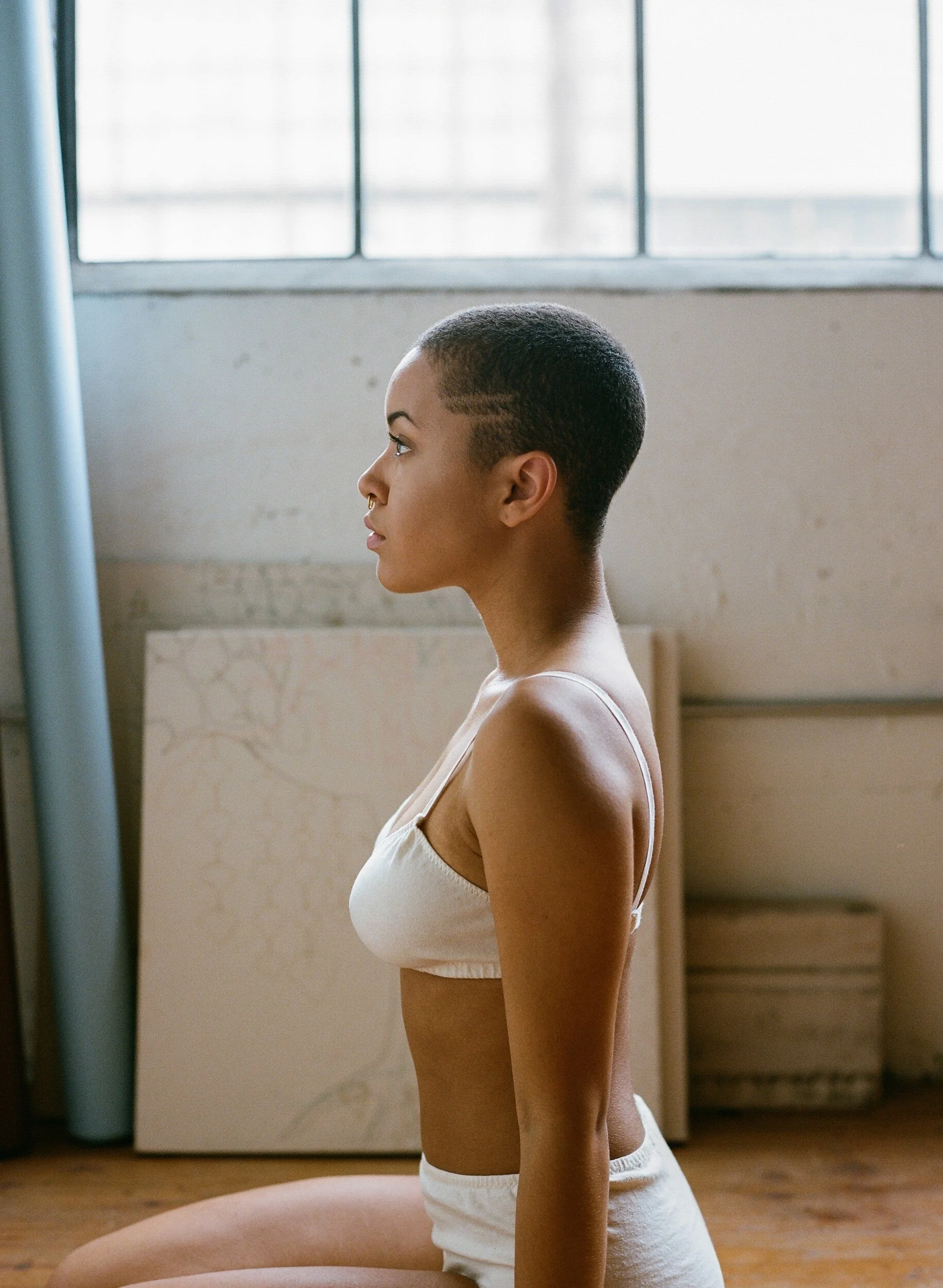 A model with closely cropped hair sits in profile wearing cream underwear beside a large window.