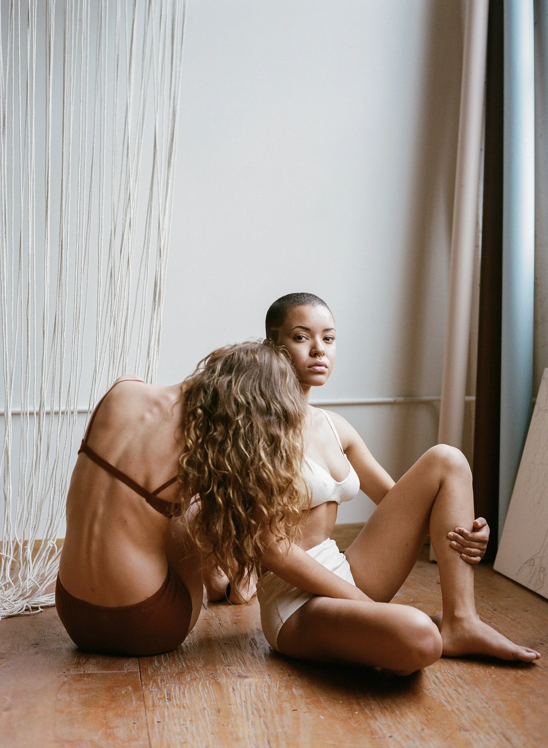Two models in rust and cream underwear sit on a wooden floor, one leaning their head against the other’s shoulder.