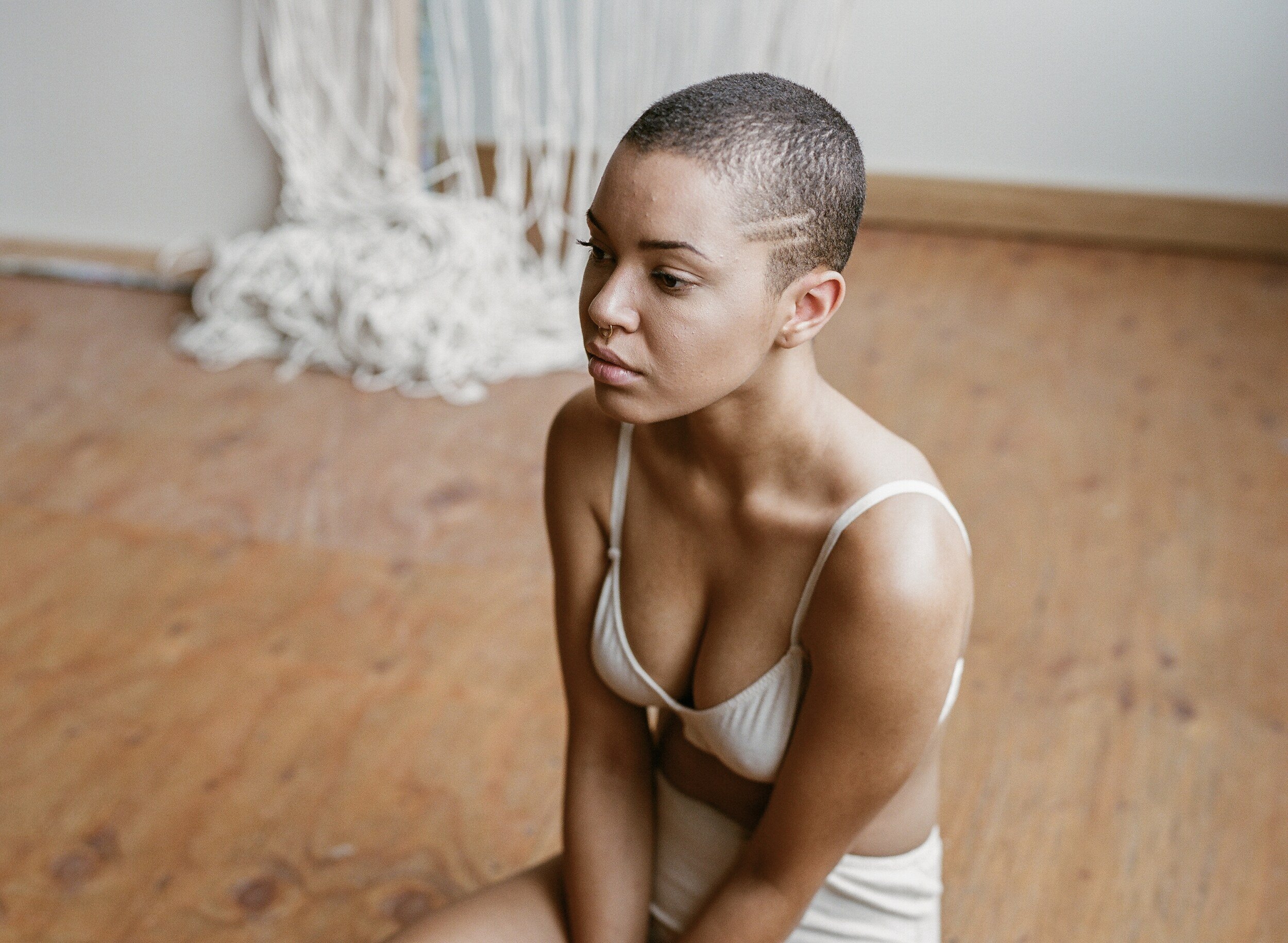 A model in cream underwear sits on a wooden floor, seen from above with their face turned to one side.