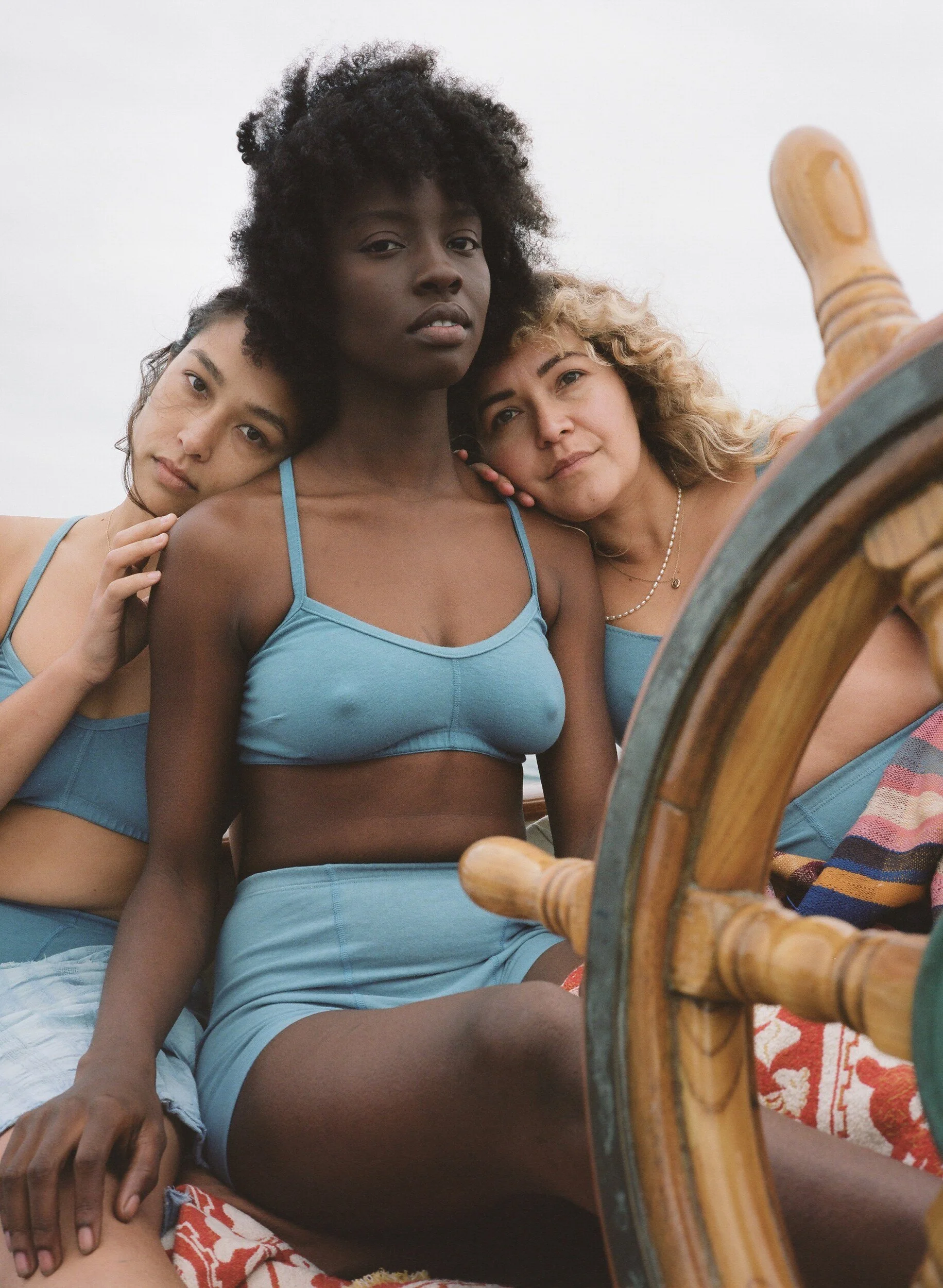 Three models in blue underwear lean together beside a wooden ship’s wheel.