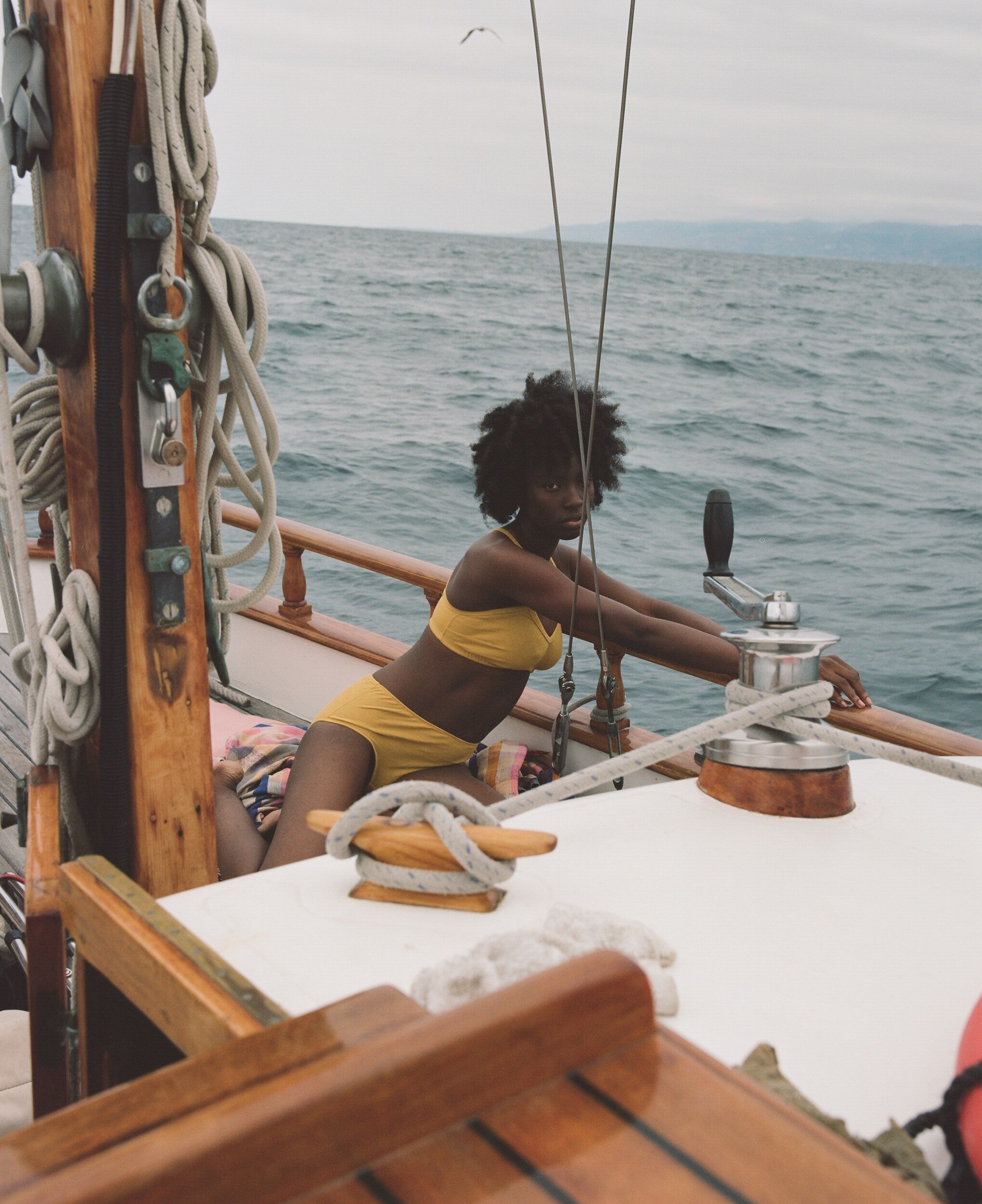 A model in yellow underwear leans against the sailboat’s wooden railing, with open water behind.
