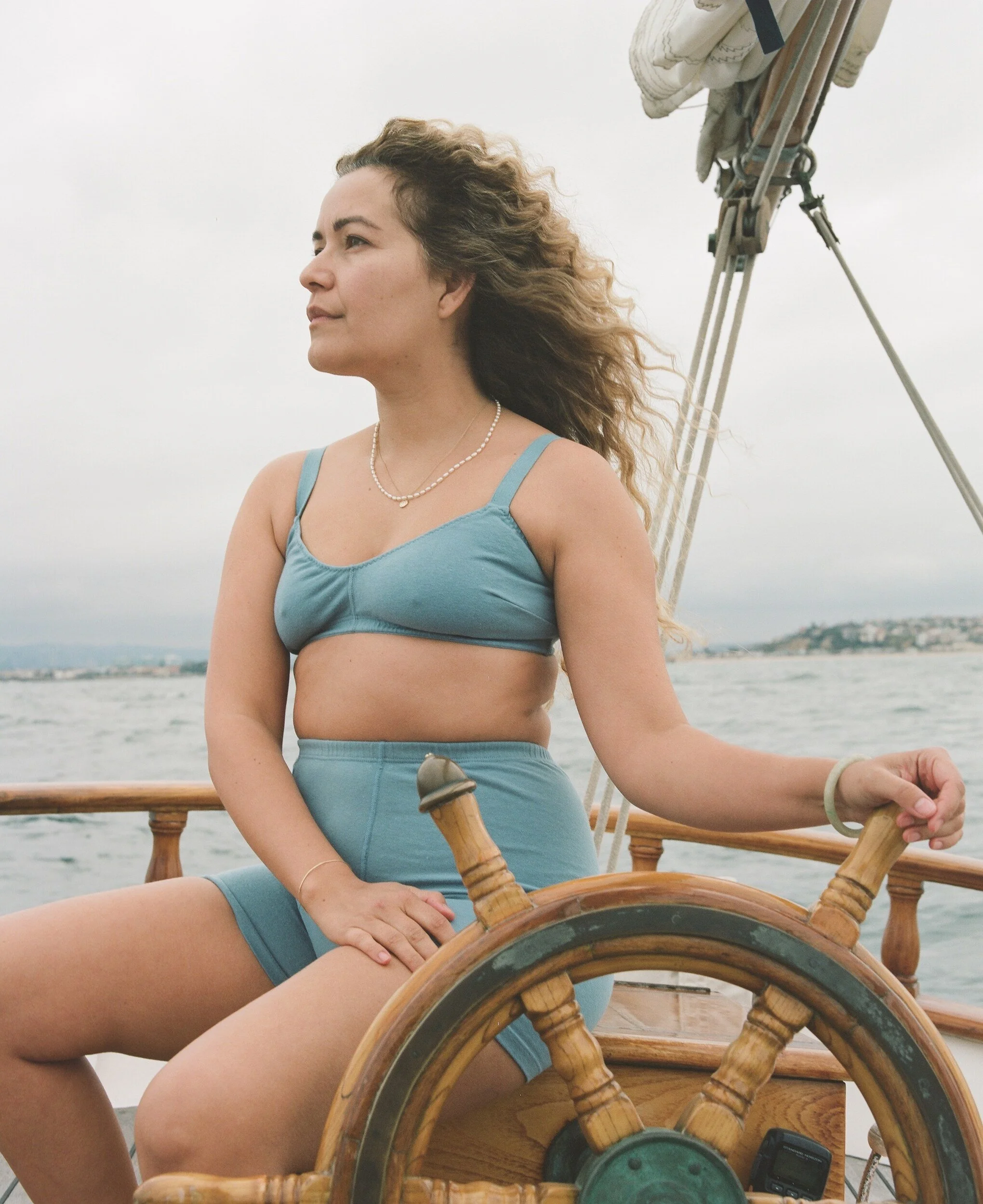 A model in a blue bra and high-waisted briefs sits at the wheel, looking out over the water.