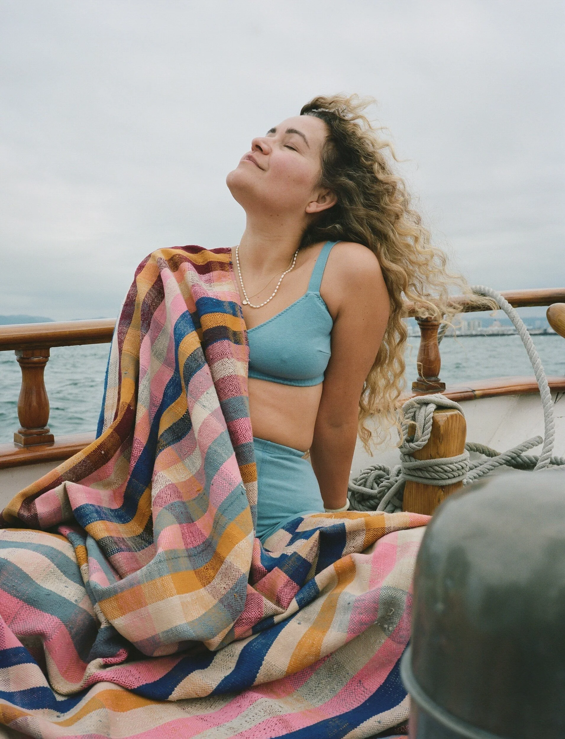A model in a blue bra tilts their face upward, wrapped in a striped blanket on the boat.