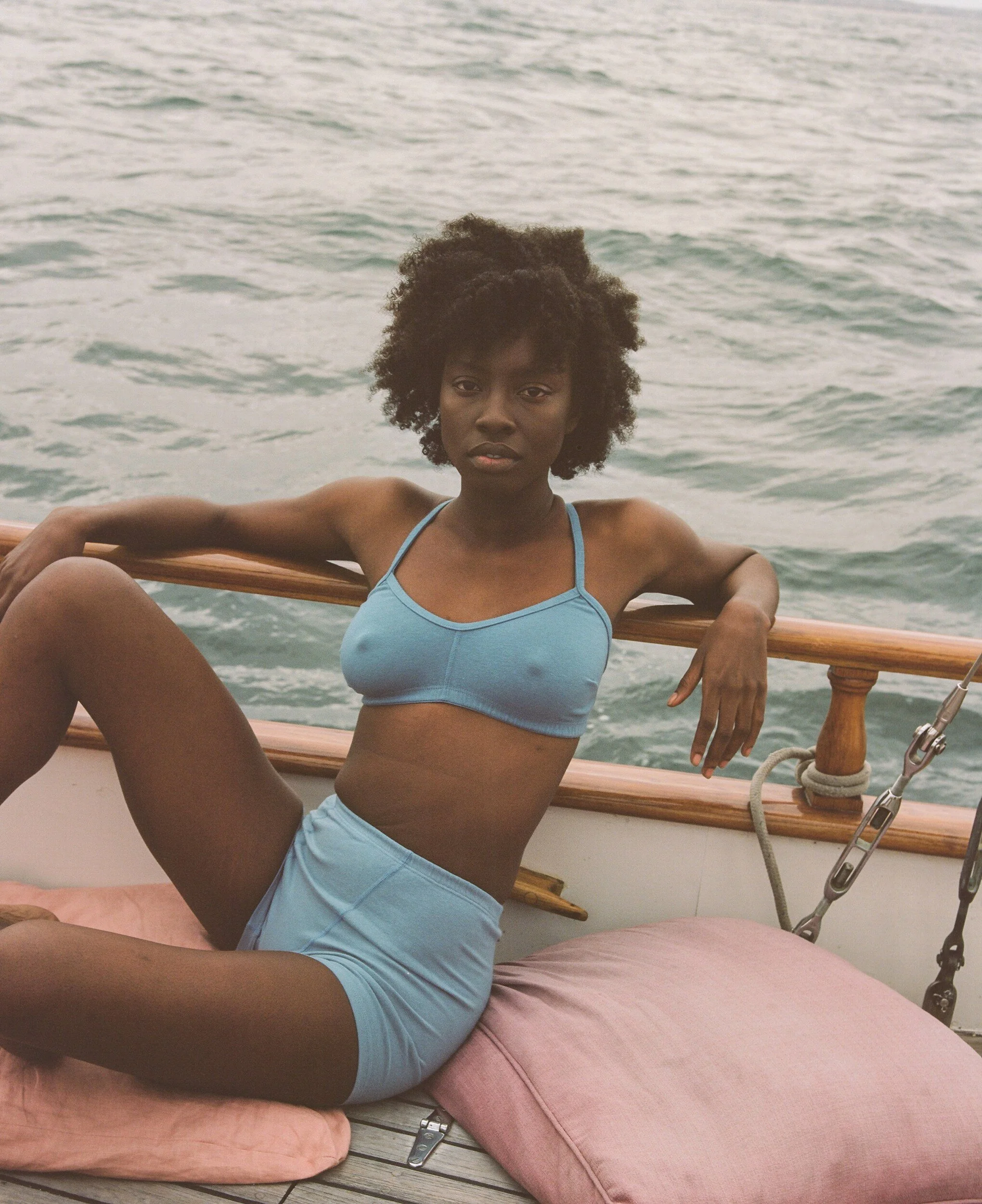 A model in blue underwear lounges on pink cushions, with one arm stretched along the boat’s railing.
