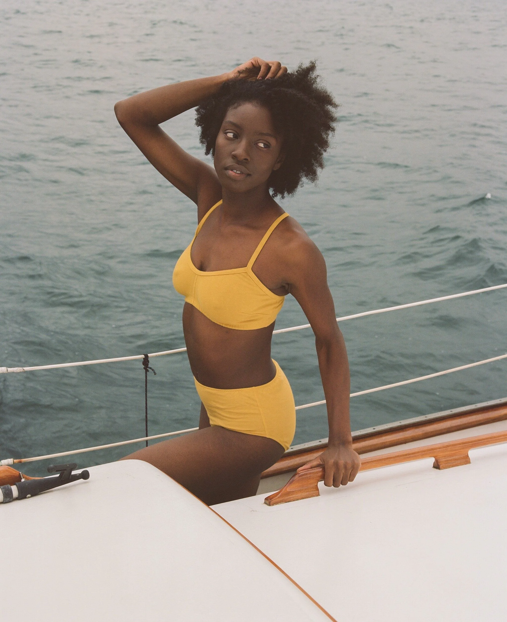 A model in a yellow bra and briefs sits at the boat’s edge with one hand in their hair.