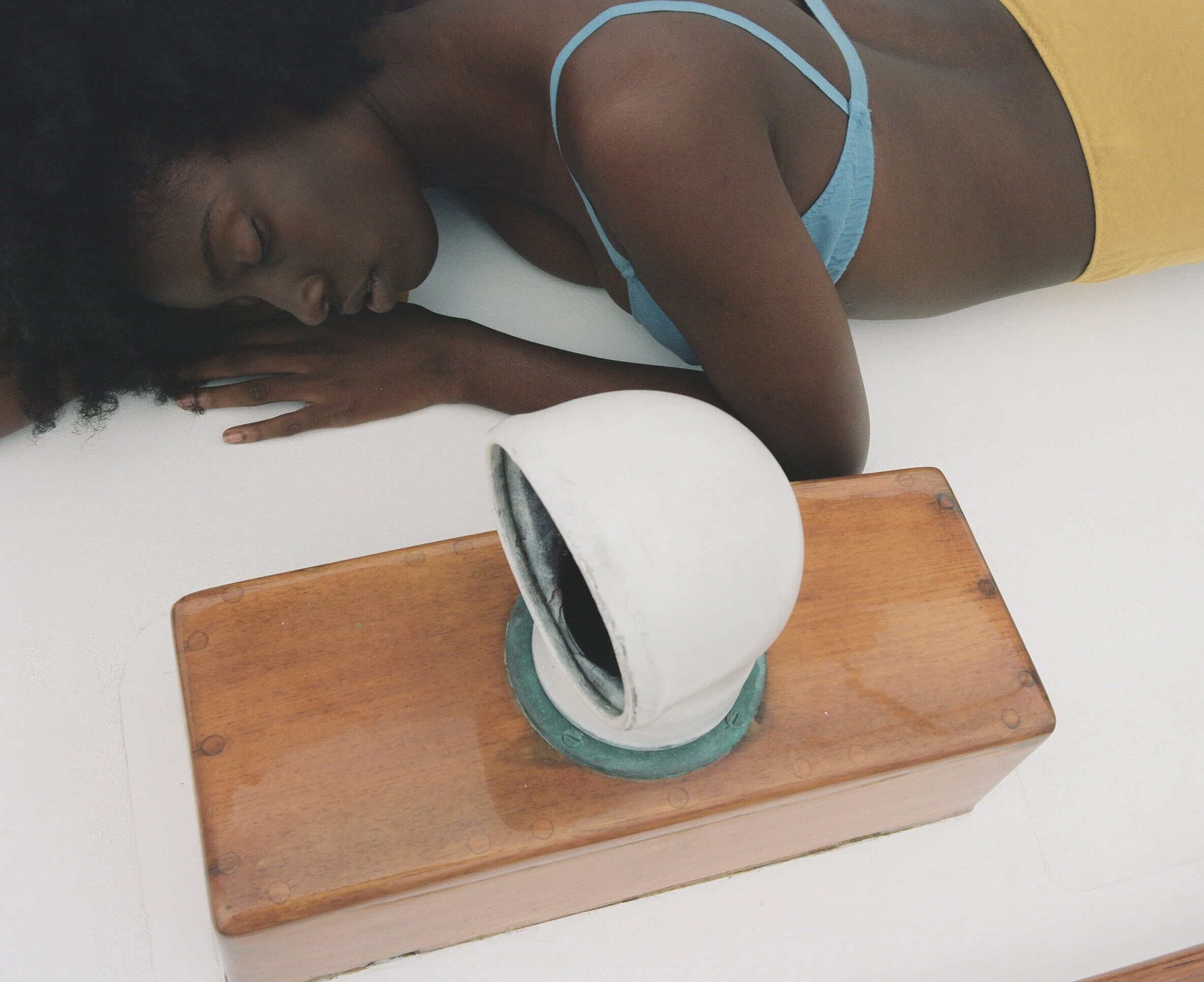 A model in a blue bra and yellow briefs rests their head on their hands beside a deck vent.