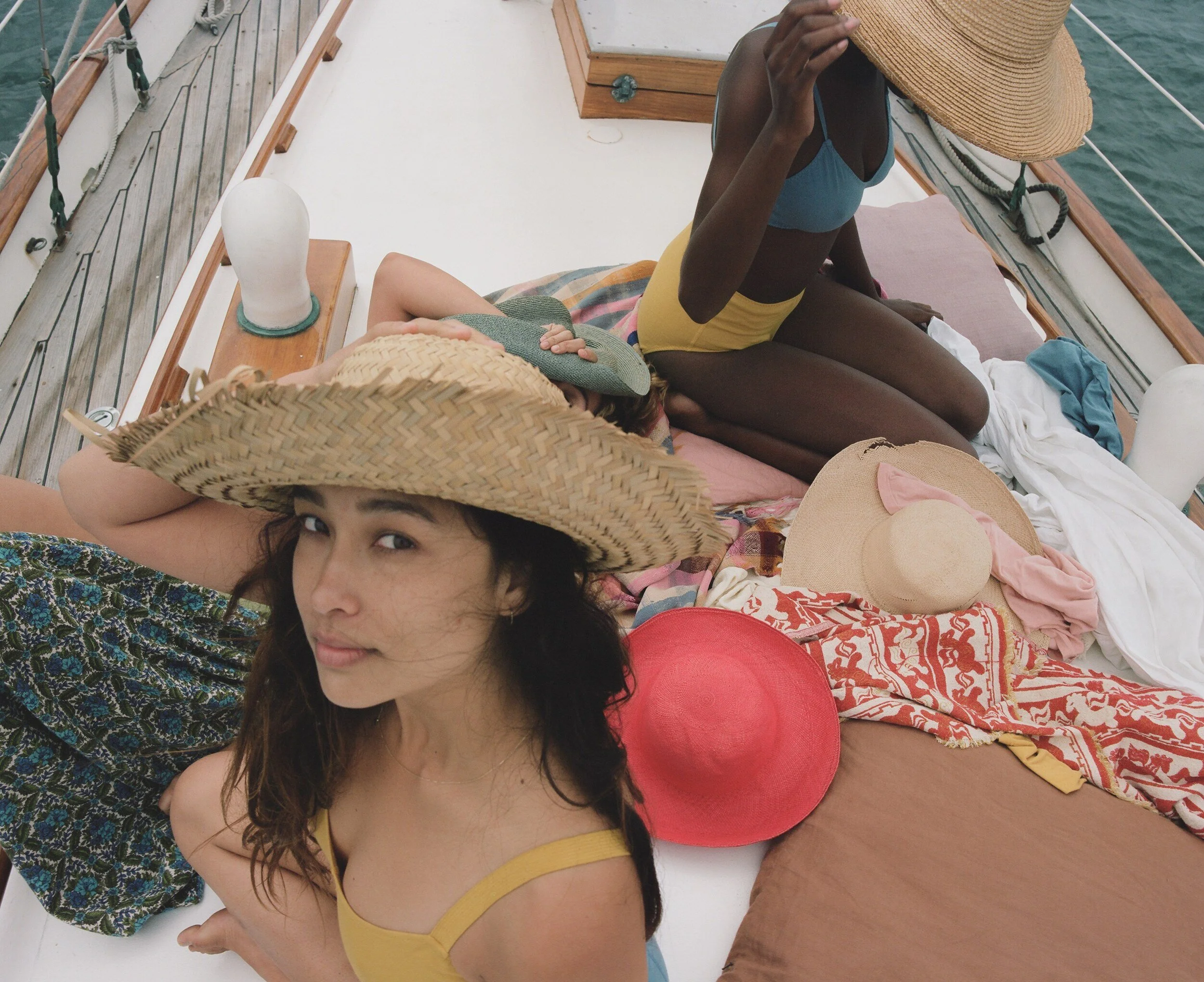 A model in a yellow bra looks up beneath a straw hat, with two other models on the deck behind.