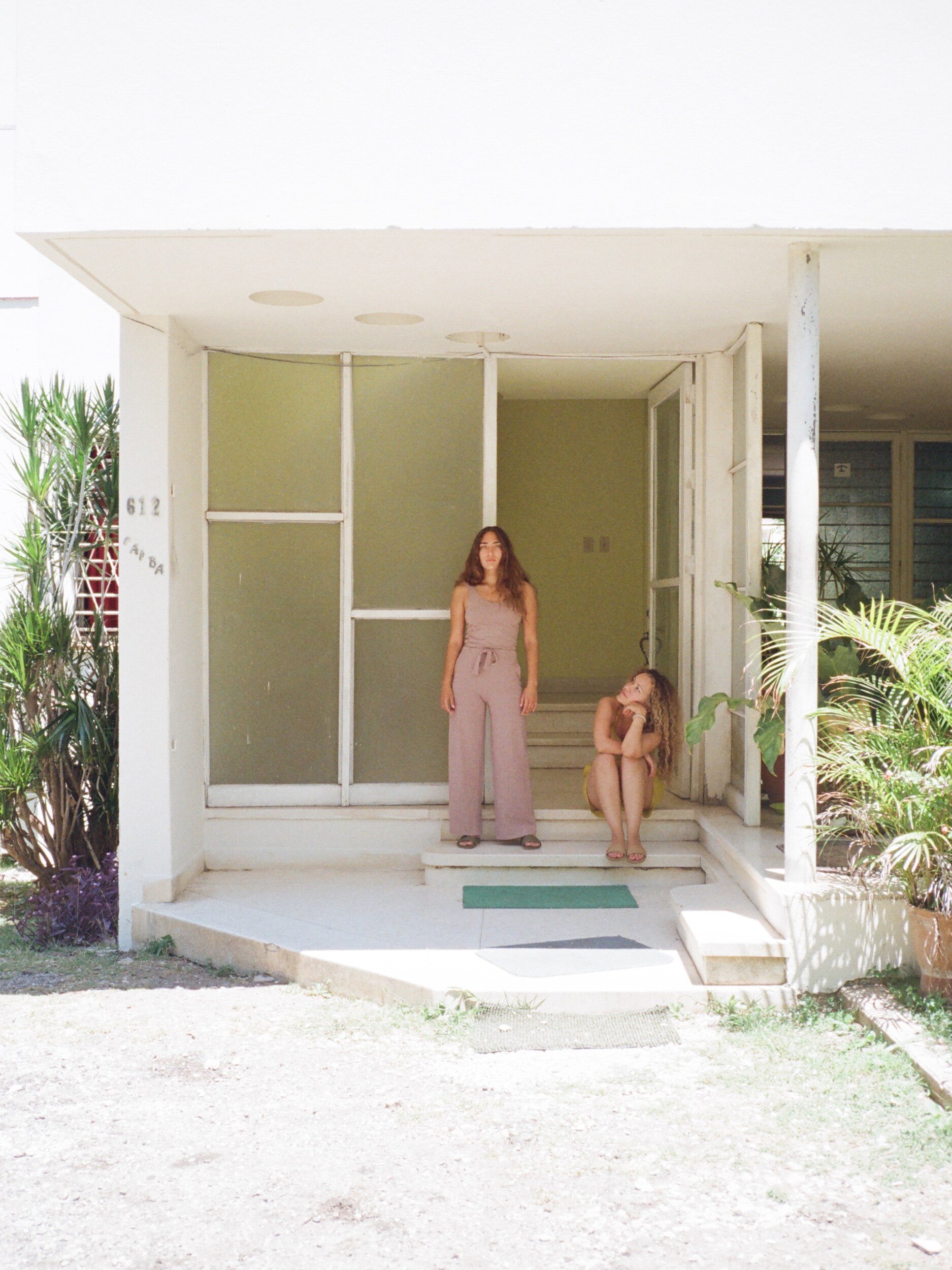 A model in a mauve tank and loose pants stands on white front steps beside another seated model.