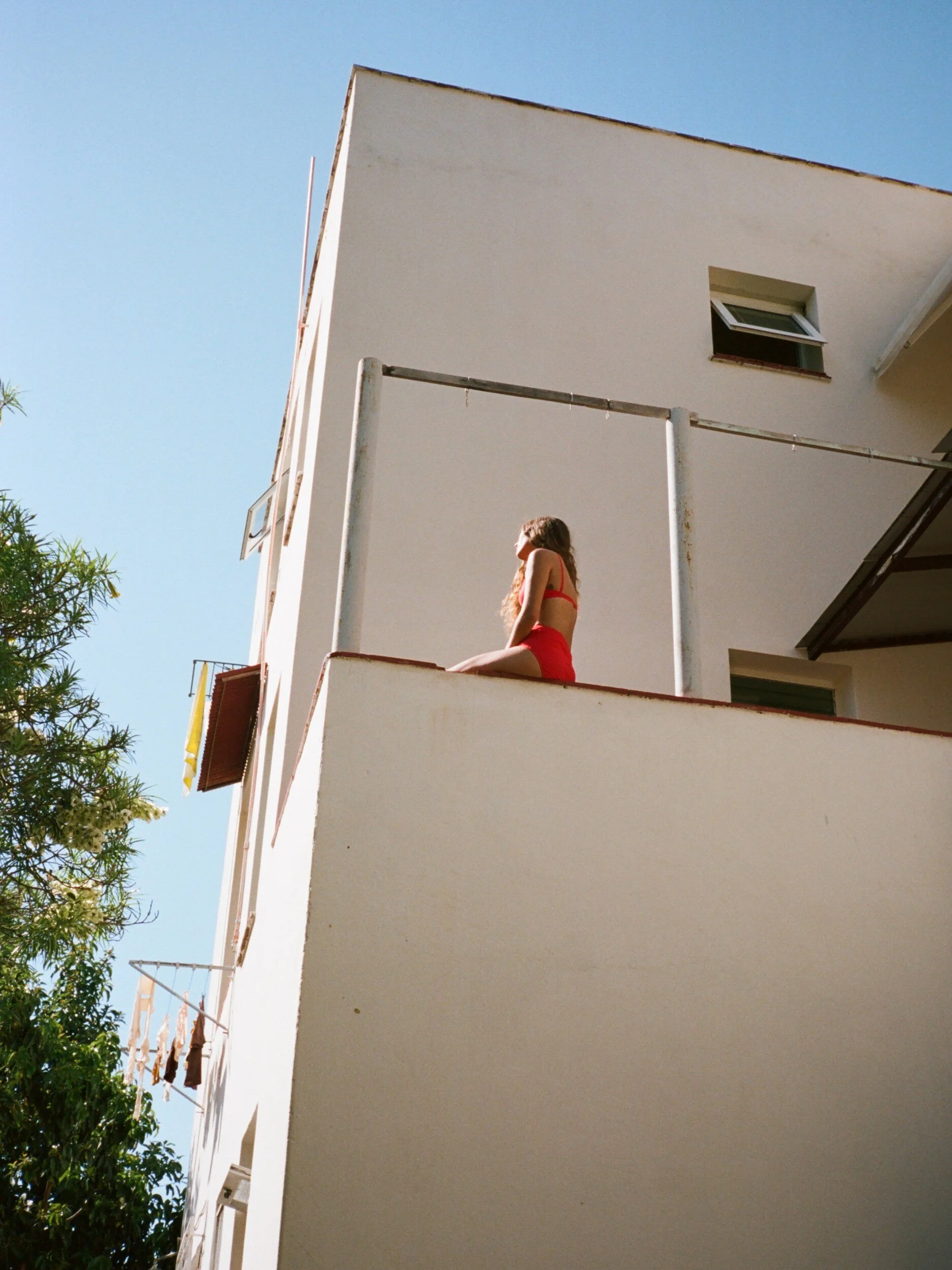 A model in red underwear sits on a white balcony wall beneath a clear blue sky.