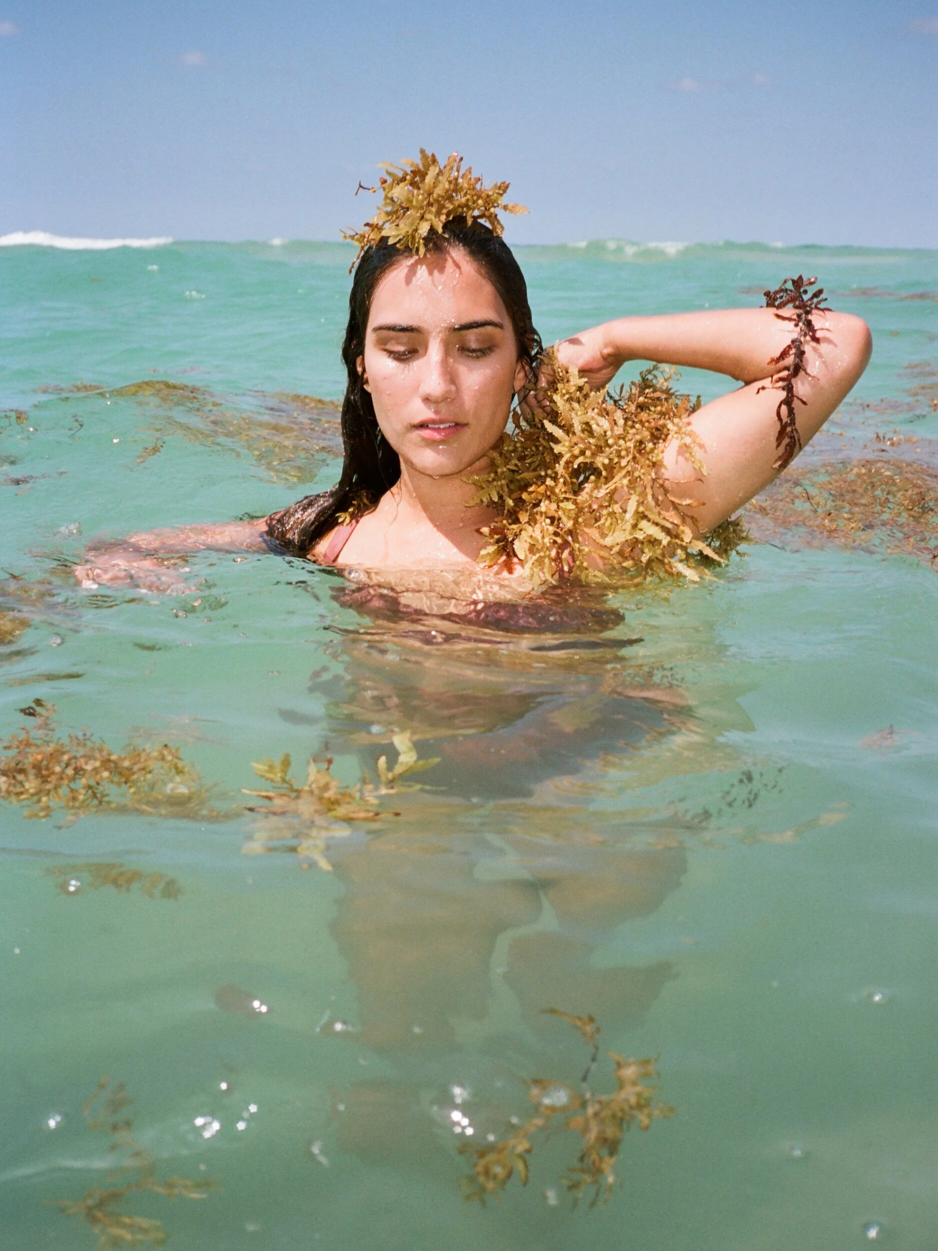 A model raises one arm in turquoise water, with seaweed piled on their head and shoulder.