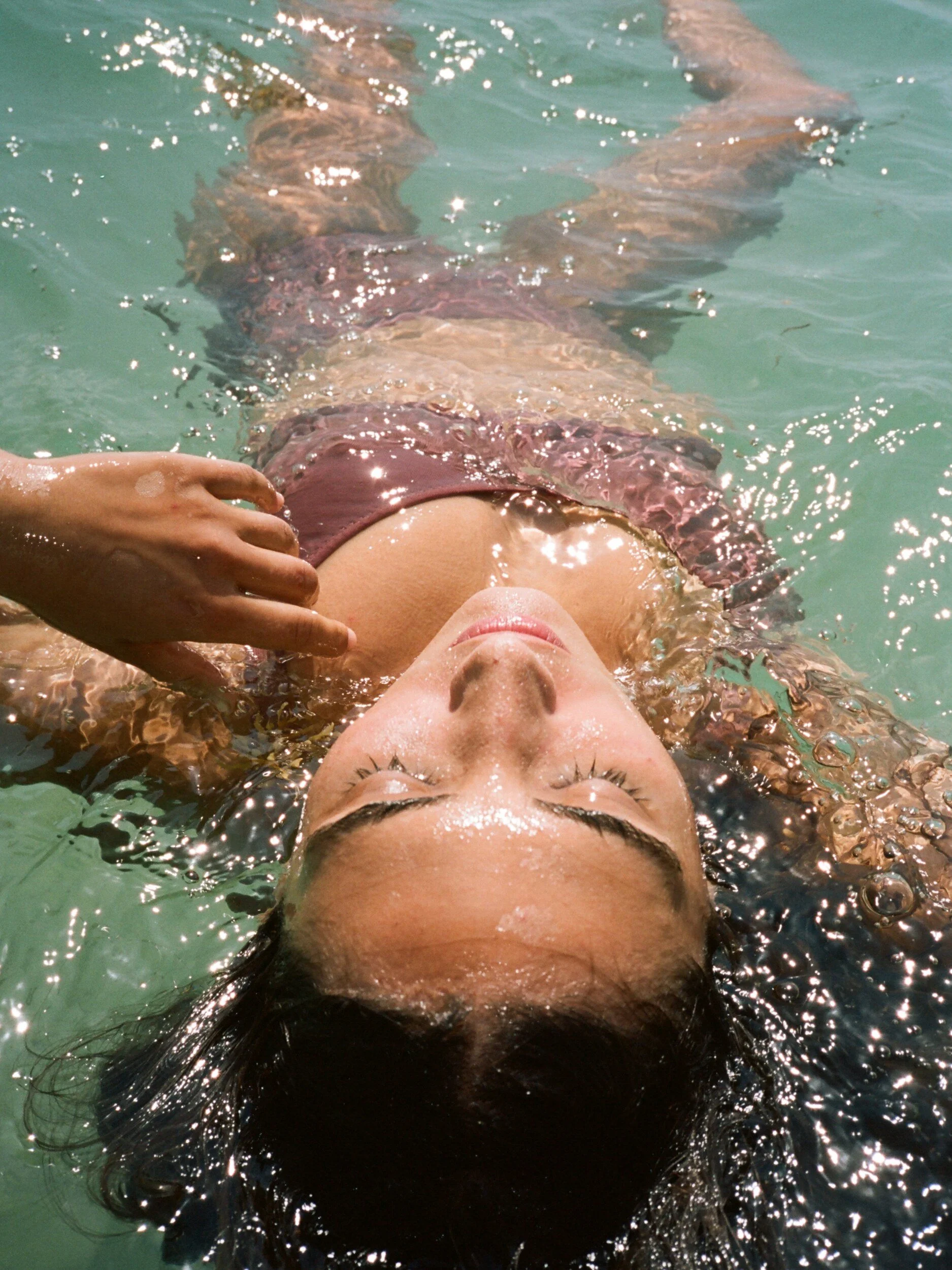 A model in mauve underwear floats on their back, eyes closed and hair spreading through the turquoise water.