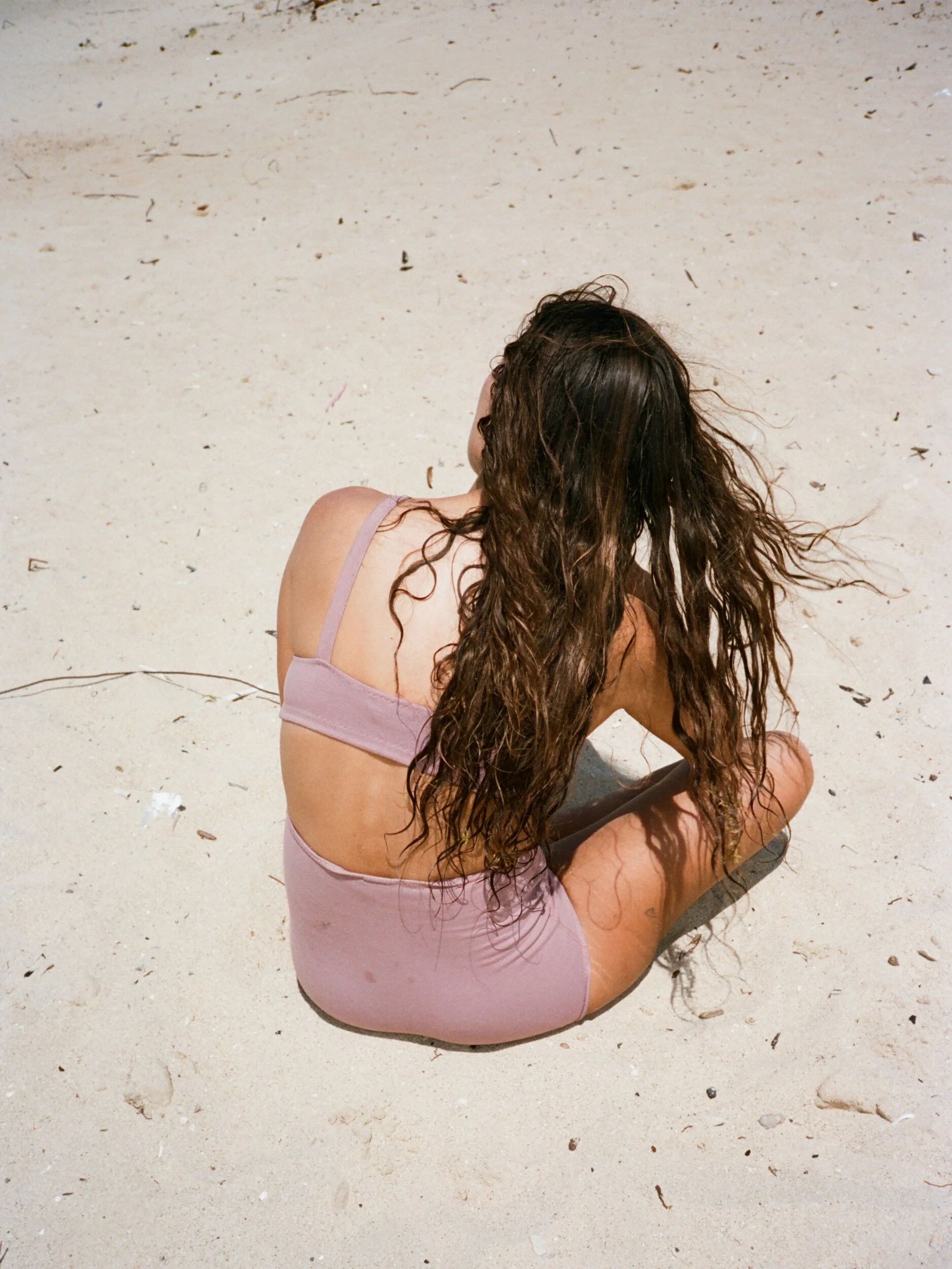 Seen from behind, a model in mauve underwear sits on pale sand with long hair down their back.