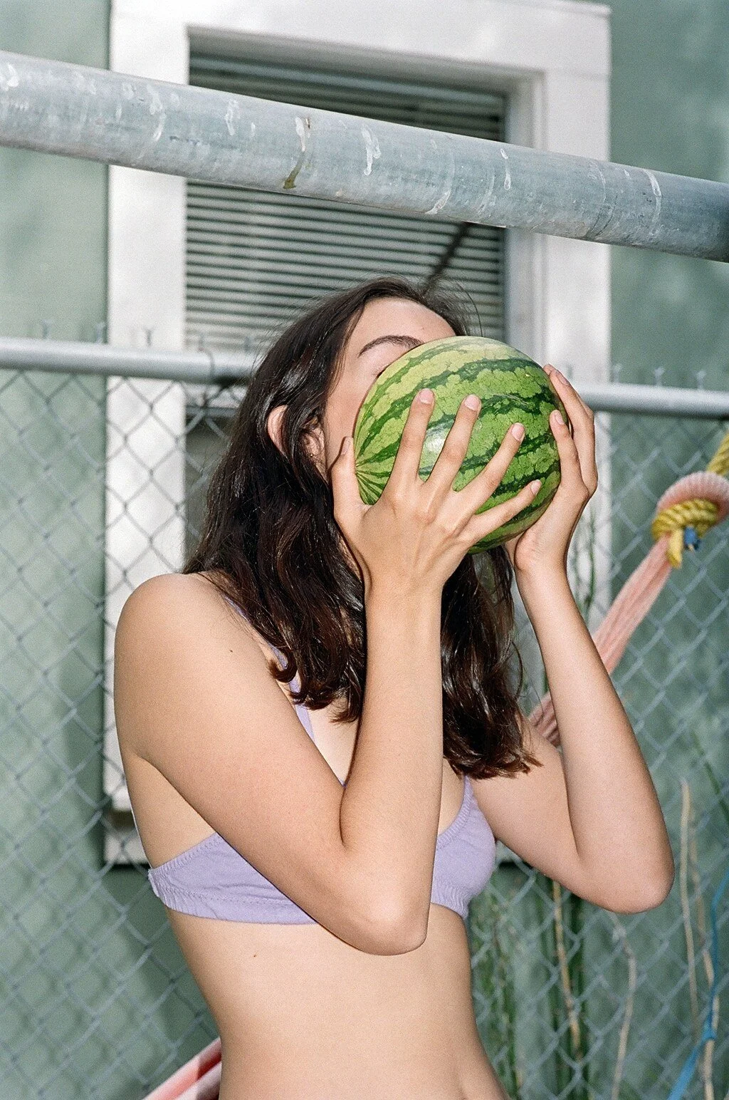 A model in a lilac bra holds a small watermelon in front of their face beside a chain-link fence.
