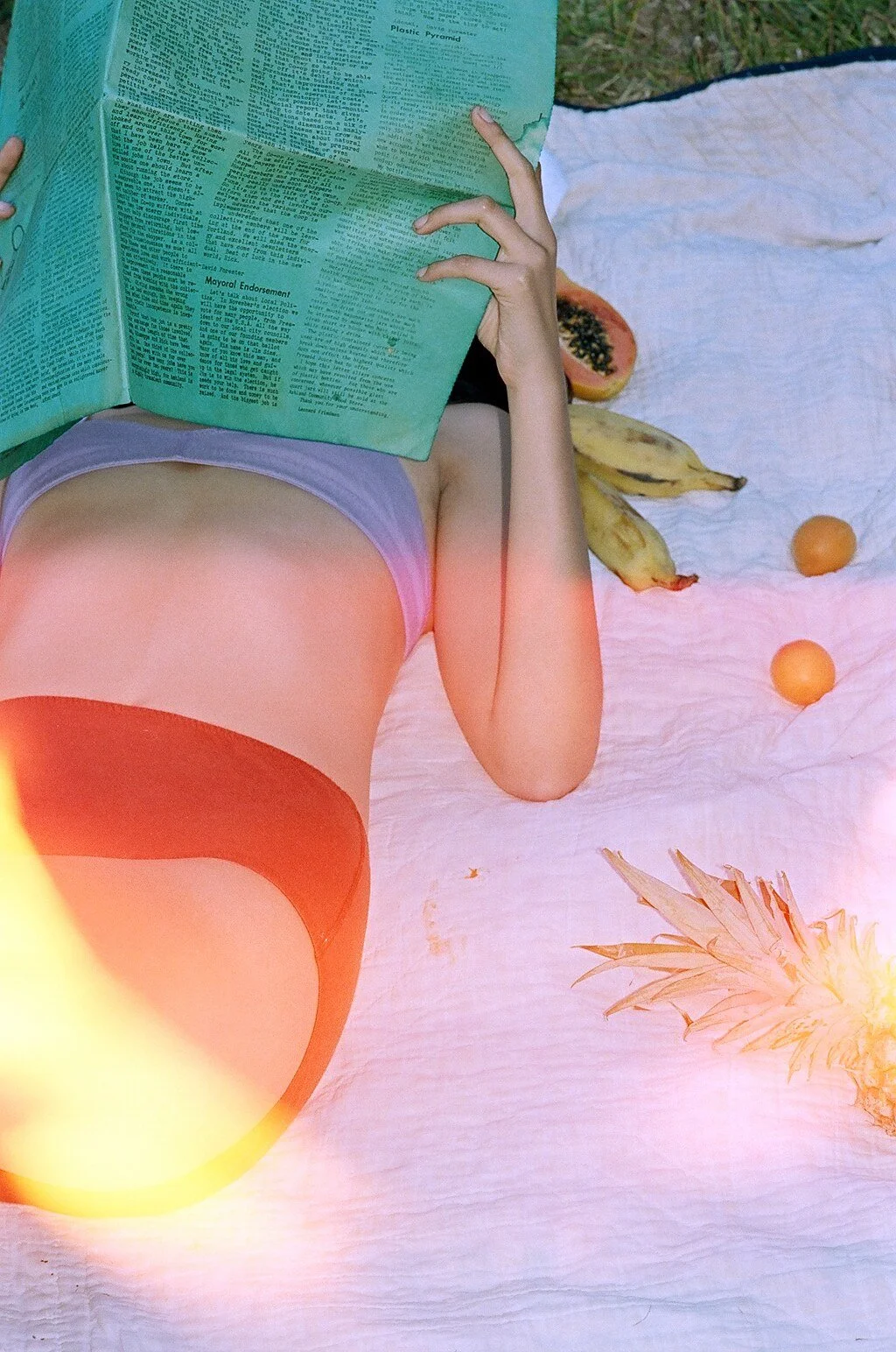 A model lies among fruit on a white blanket, with a green printed sheet covering their face and orange light across their underwear.