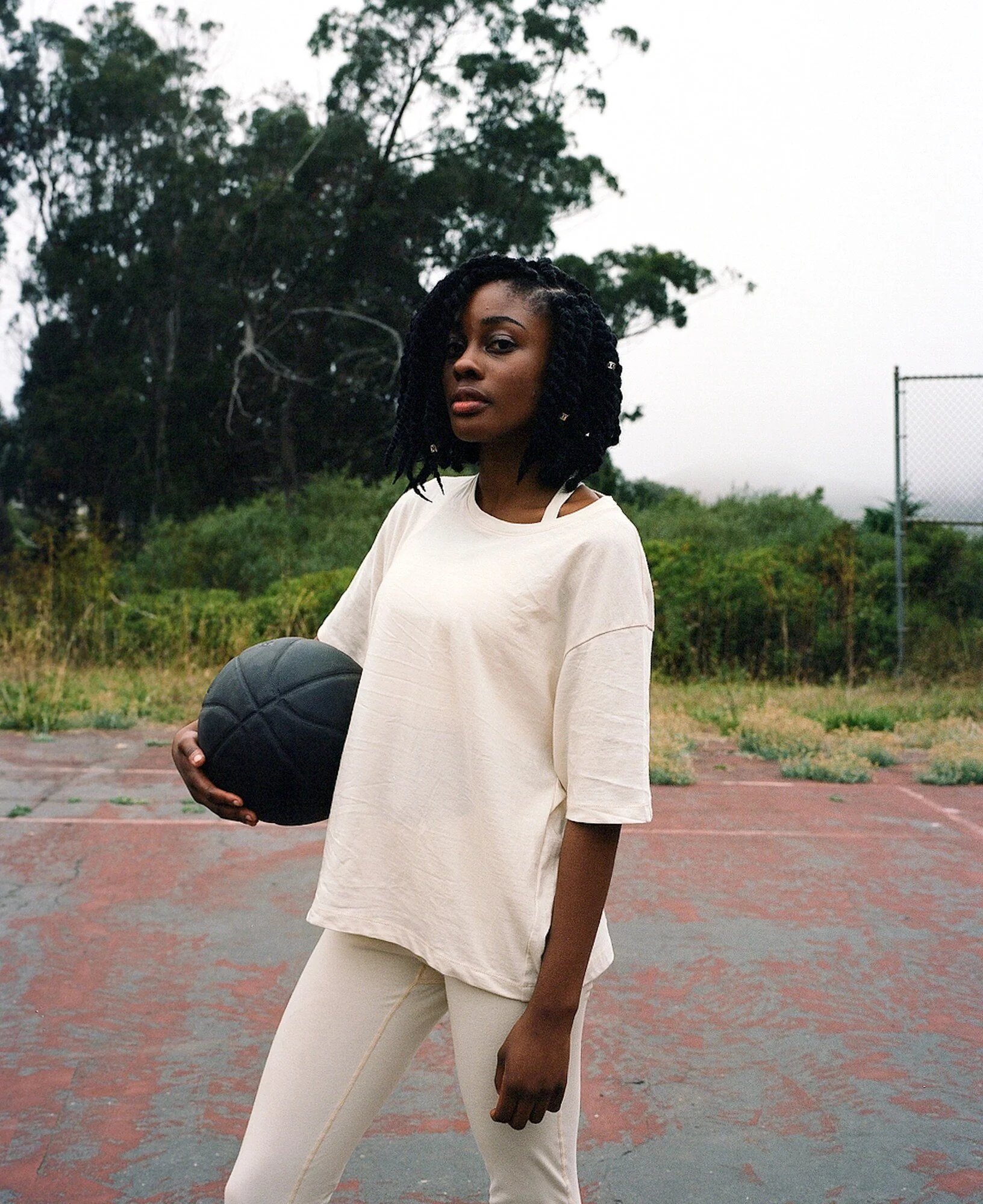 A model in a cream T-shirt and leggings holds a dark basketball on a worn outdoor court.