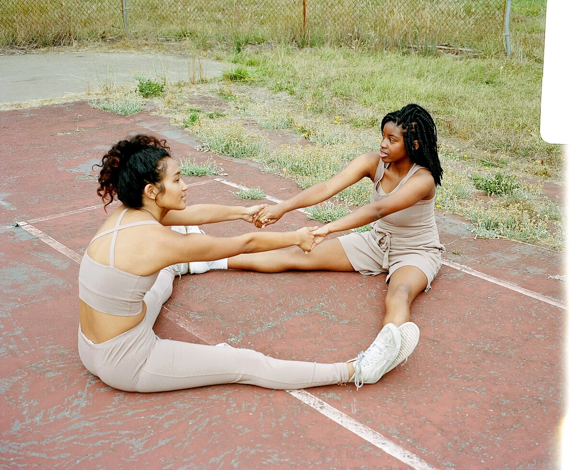 Two models in mauve sportswear sit facing each other with legs apart, holding hands as they stretch.