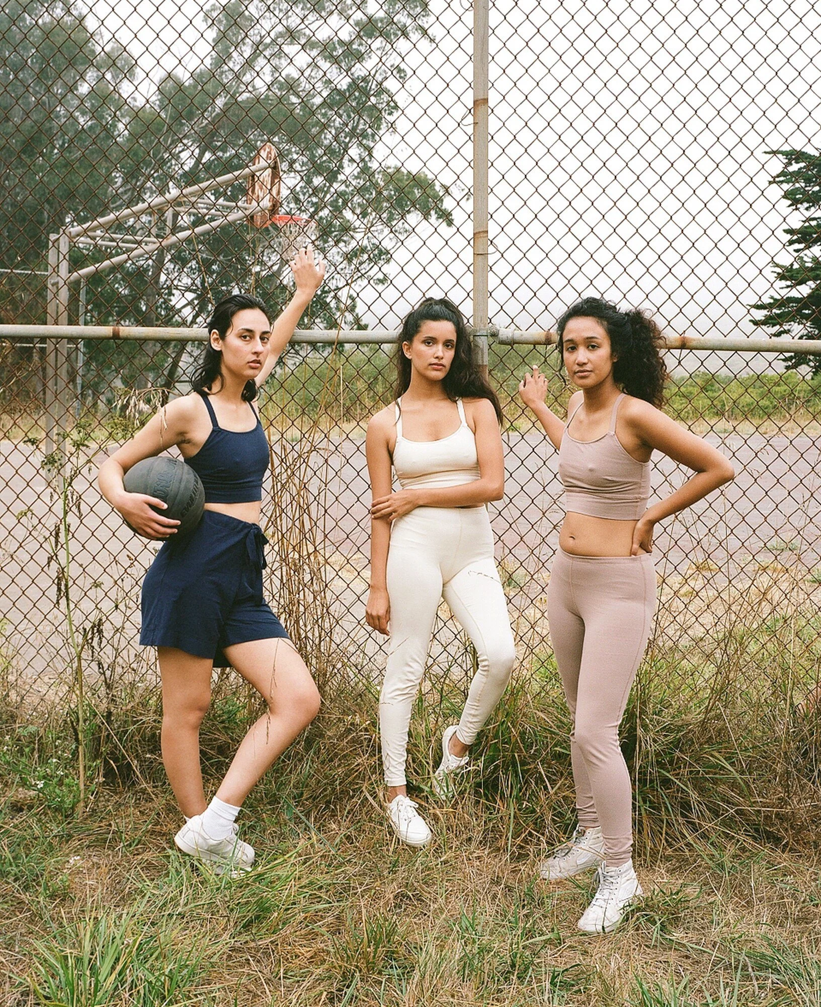 Three models in navy, cream and mauve sportswear stand beside a chain-link fence, one holding a basketball.
