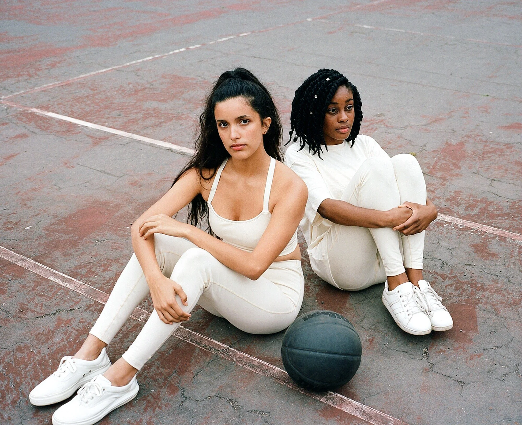 Two models in cream sportswear sit back to back on the court with a dark basketball beside them.