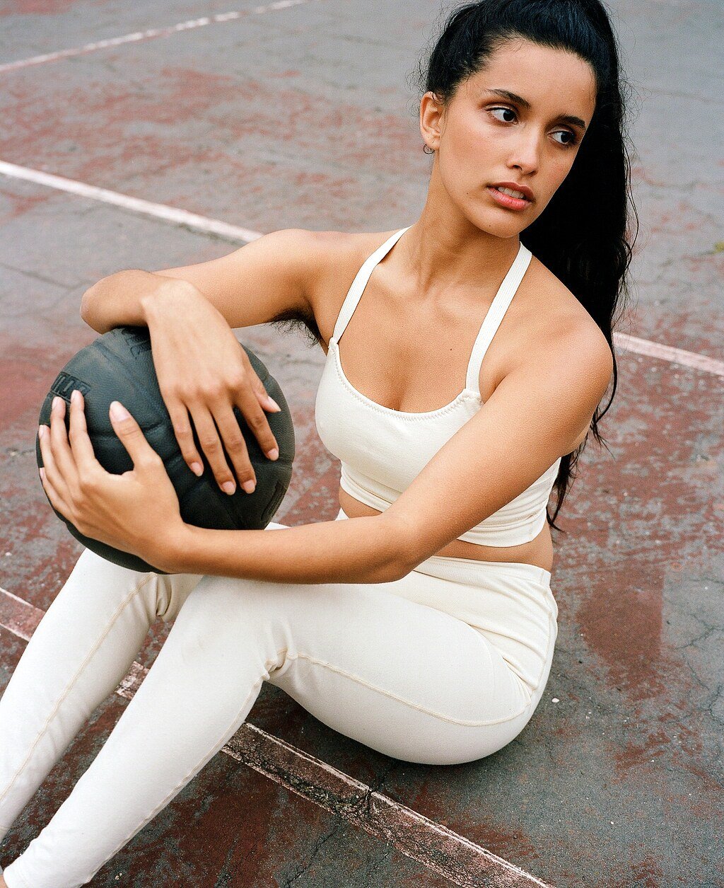 A model in a cream crop top and leggings sits on the court holding a dark basketball against one knee.