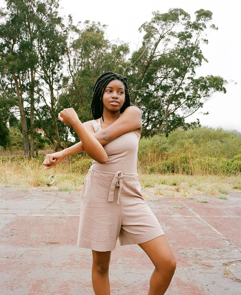A model in a mauve tank and drawstring shorts stretches one arm across their chest.