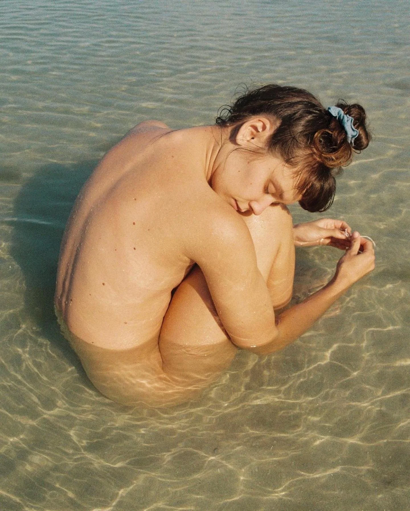 A pale blue scrunchie holds a model’s hair in a bun as they sit in clear, shallow water.