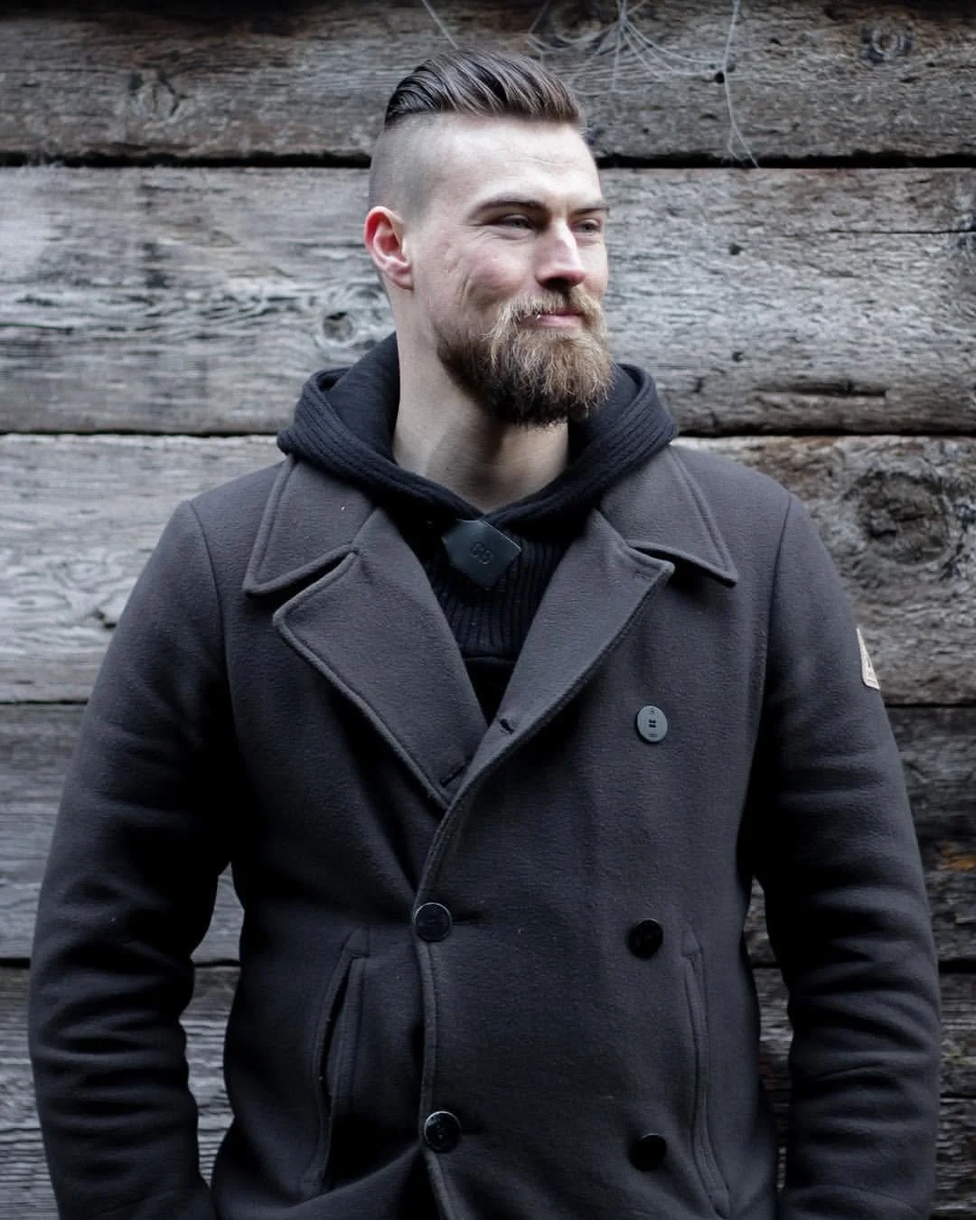 A man with a beard and styled hair is standing in front of a rustic wooden wall. He is wearing a dark gray wool coat over a black hoodie.