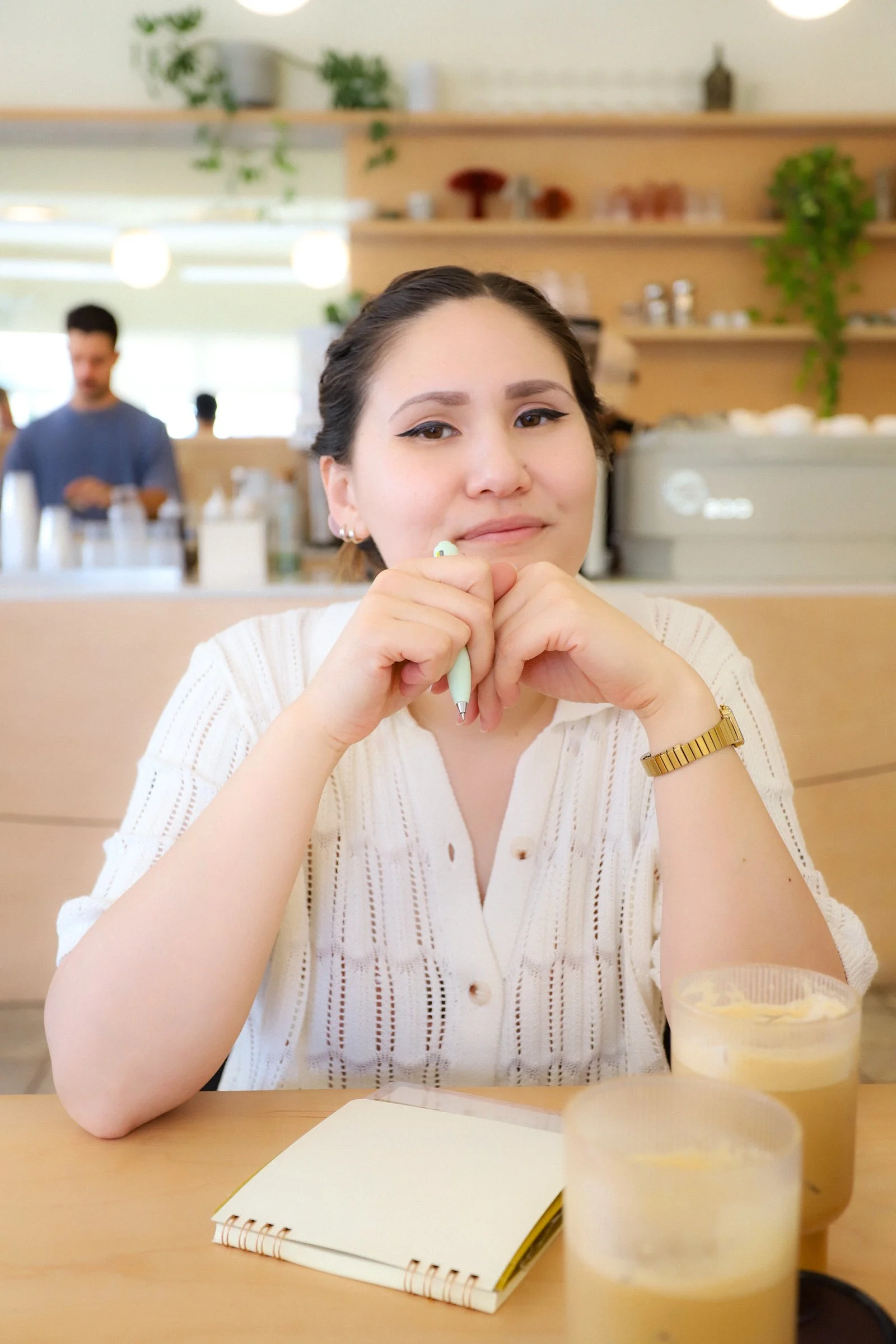 A woman sitting at a table in a cafe, holding a pen and smiling, with a notebook and two drinks in front of her.