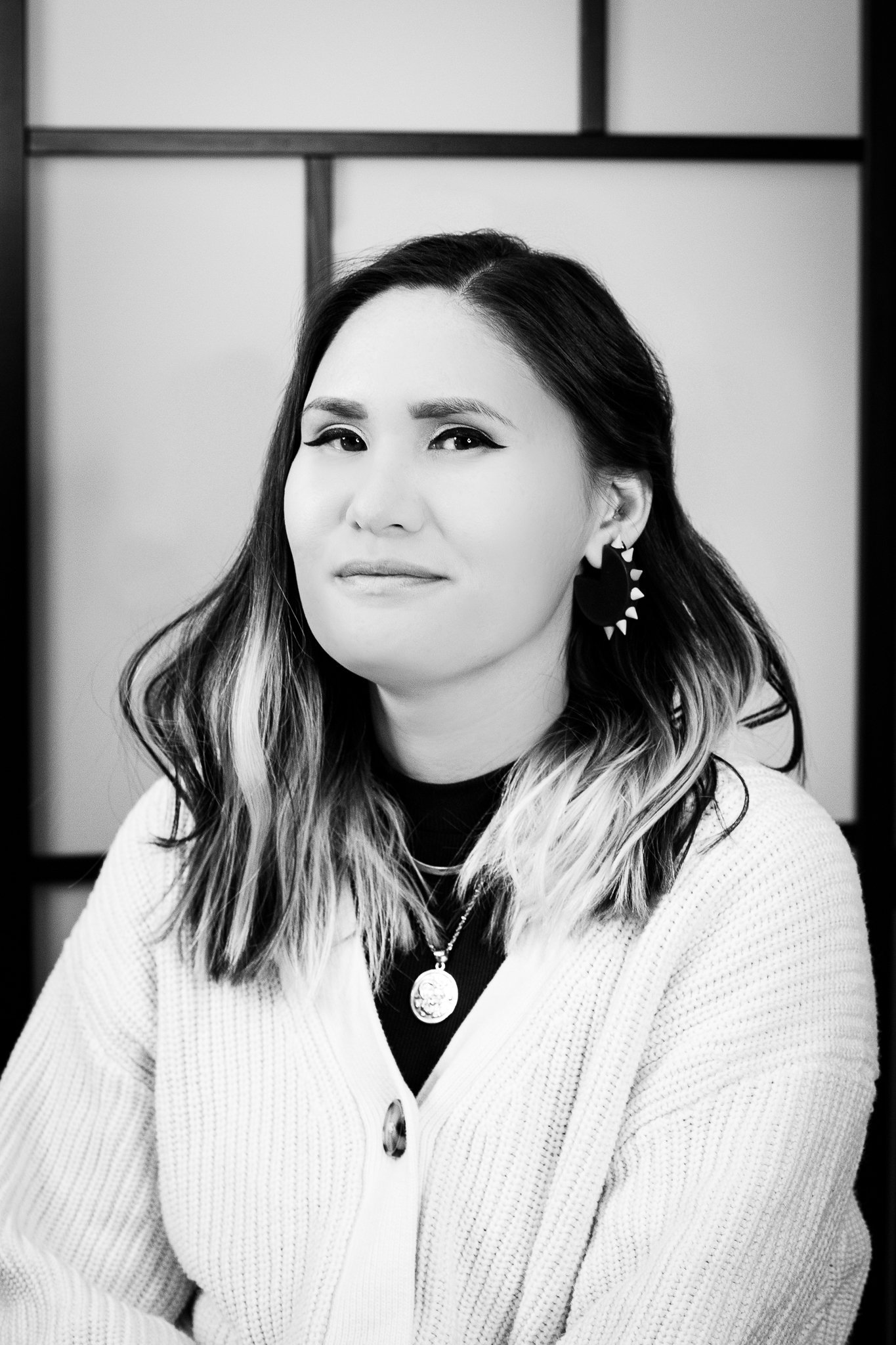 A woman with long hair wearing a white knitted cardigan, a black top, and heart-shaped earrings, posing against a plain background.