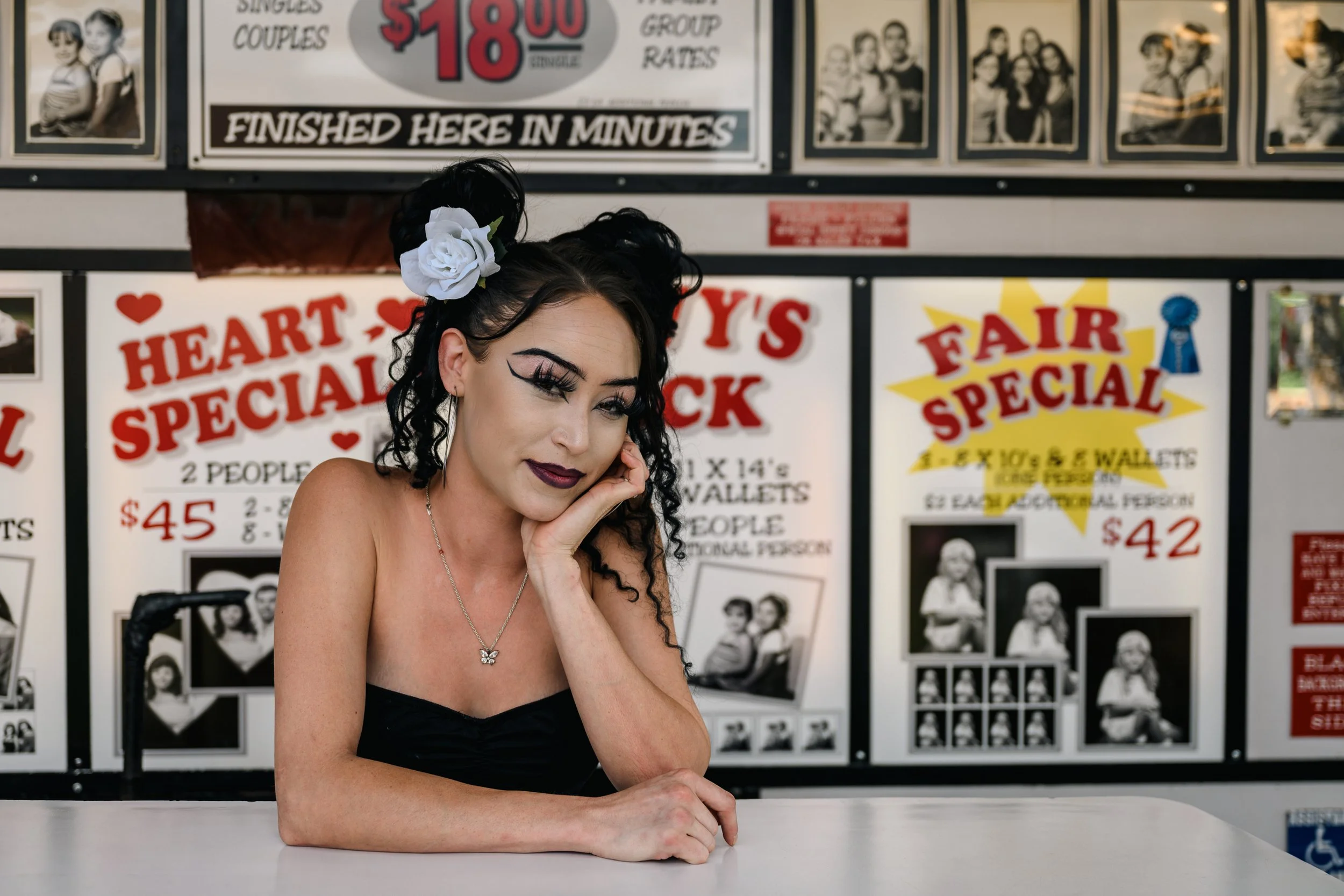A woman with dark curly hair, makeup, and a white flower in her hair, resting her head on her hand at a fairground booth with signs advertising prizes and prices in the background.