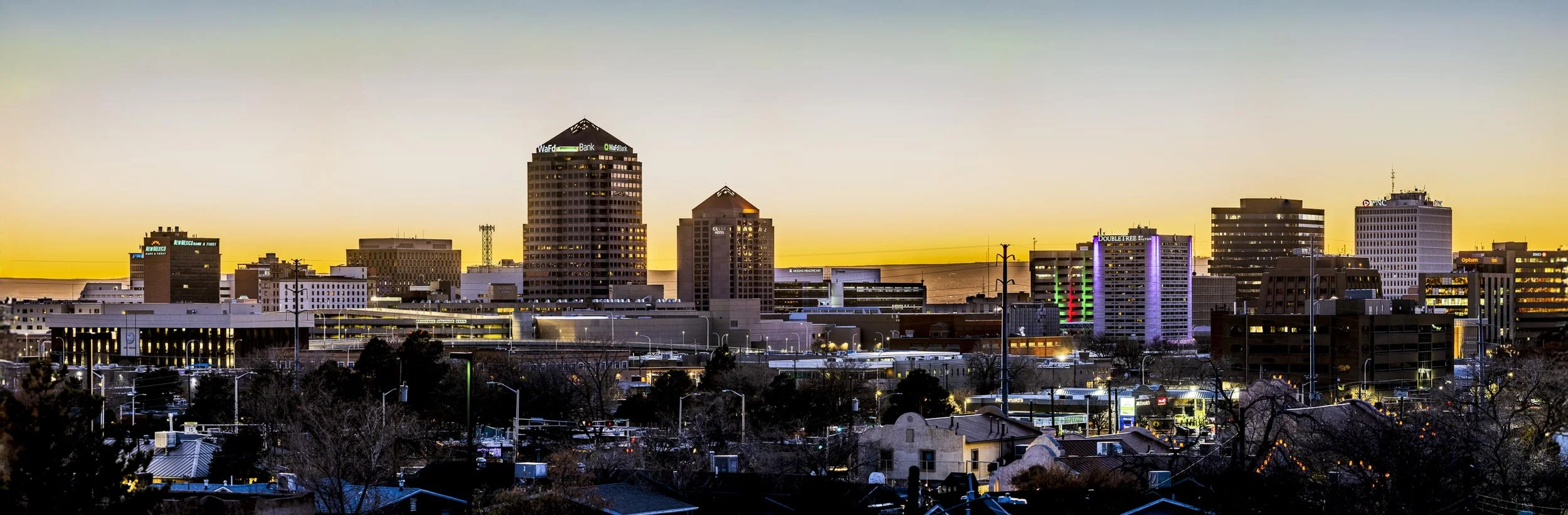 City skyline at sunset with tall buildings, some with colorful lights, and a foreground of houses and trees.
