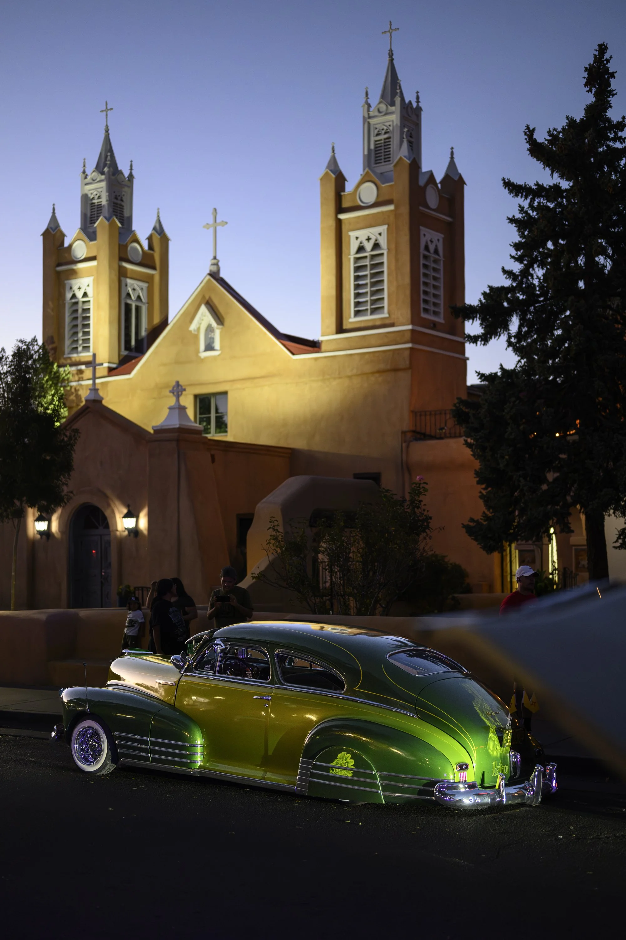 A vintage green and black car parked outside a church with tall towers, during dusk or evening.