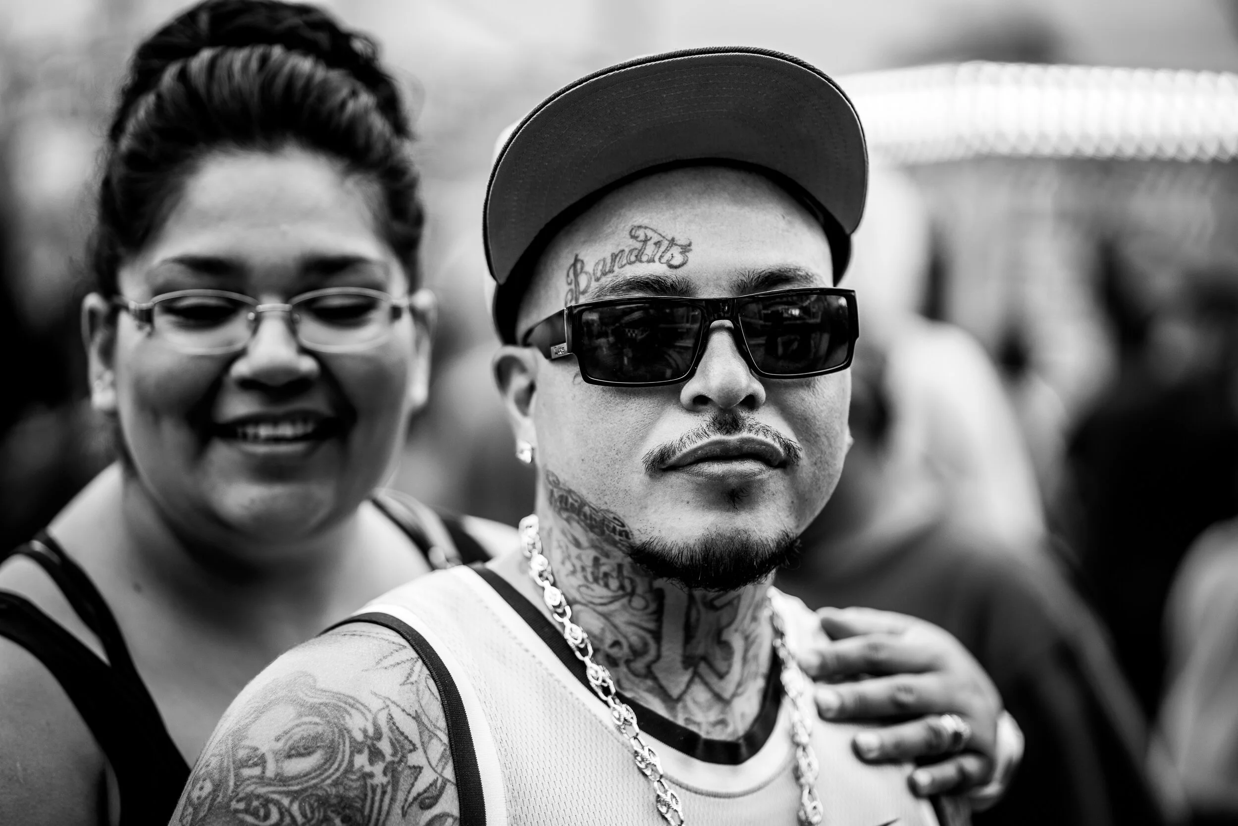 A black and white photo of a smiling woman with glasses and a man with tattoos, sunglasses, a cap, and a chain around his neck, at an outdoor event.