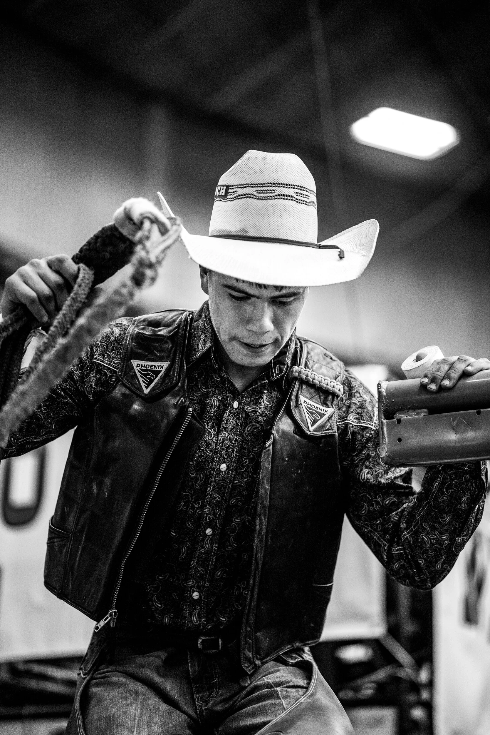 A young man dressed as a cowboy, wearing a cowboy hat and a leather vest with patches, is holding a rope in one hand and a metal object in the other, looking downward in an indoor setting.