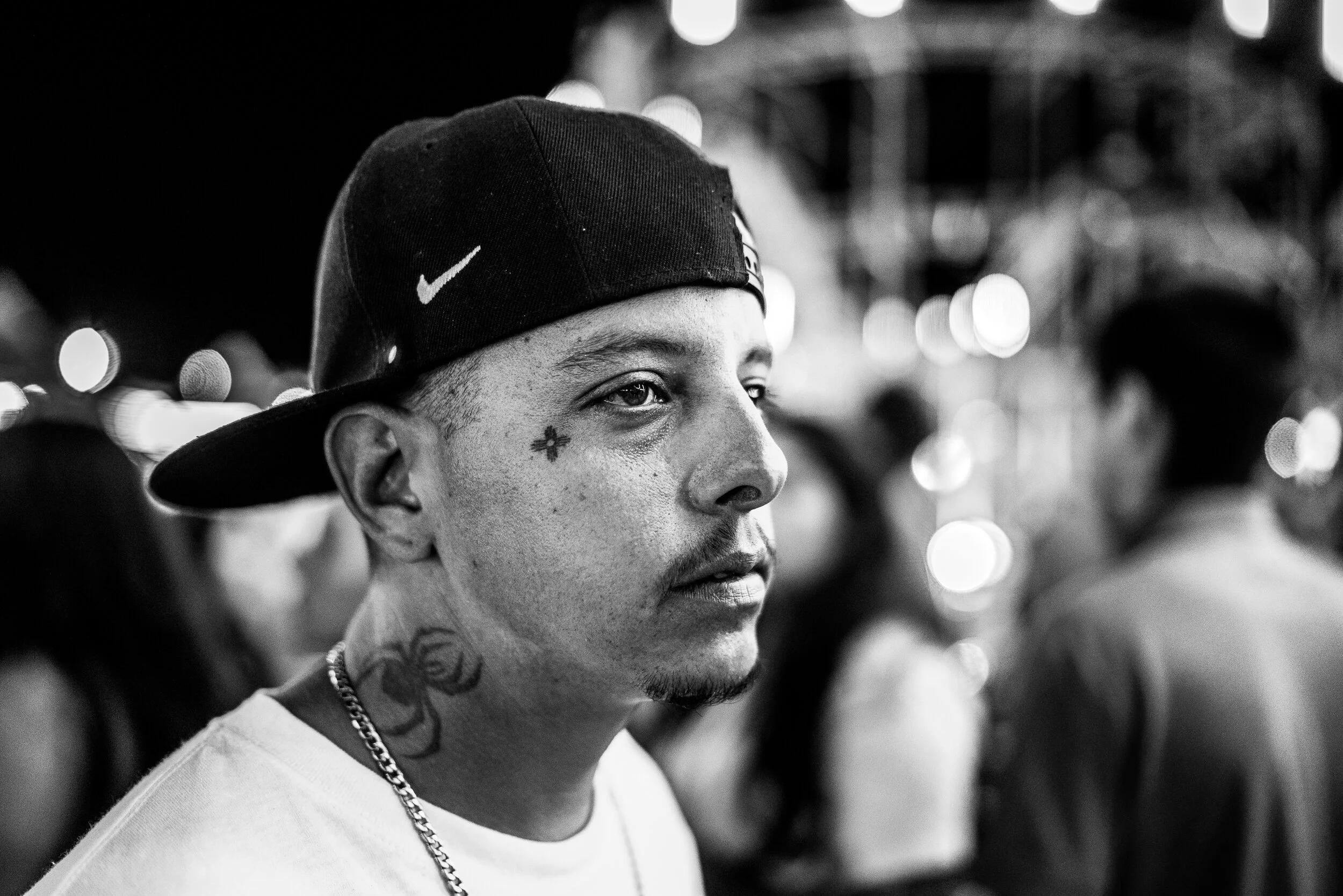 A young man with tattoos on his neck and face, wearing a backward baseball cap and a chain, stands among a crowd at night with blurred lights in the background.