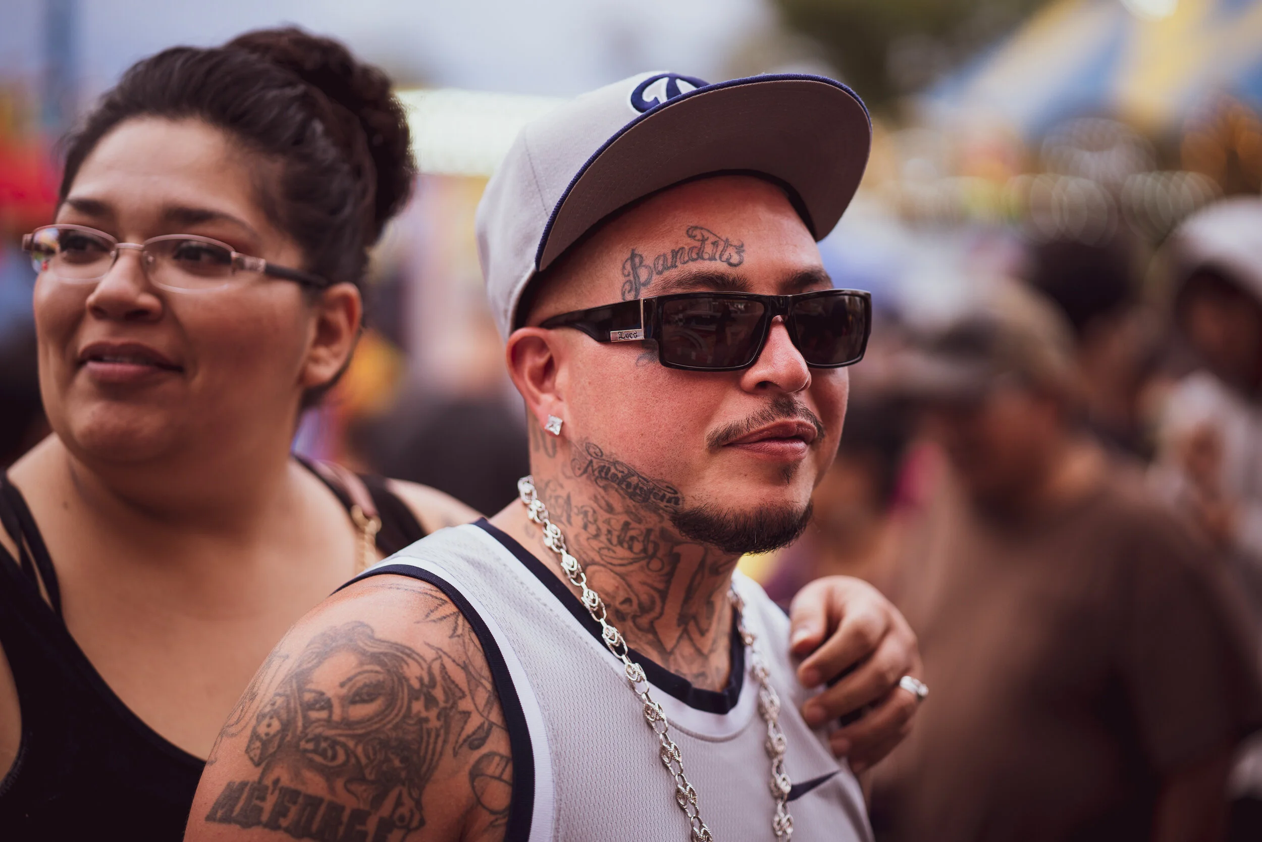 Close-up of a man with tattoos, wearing sunglasses, a baseball cap, and a chain necklace, at a crowded outdoor event.