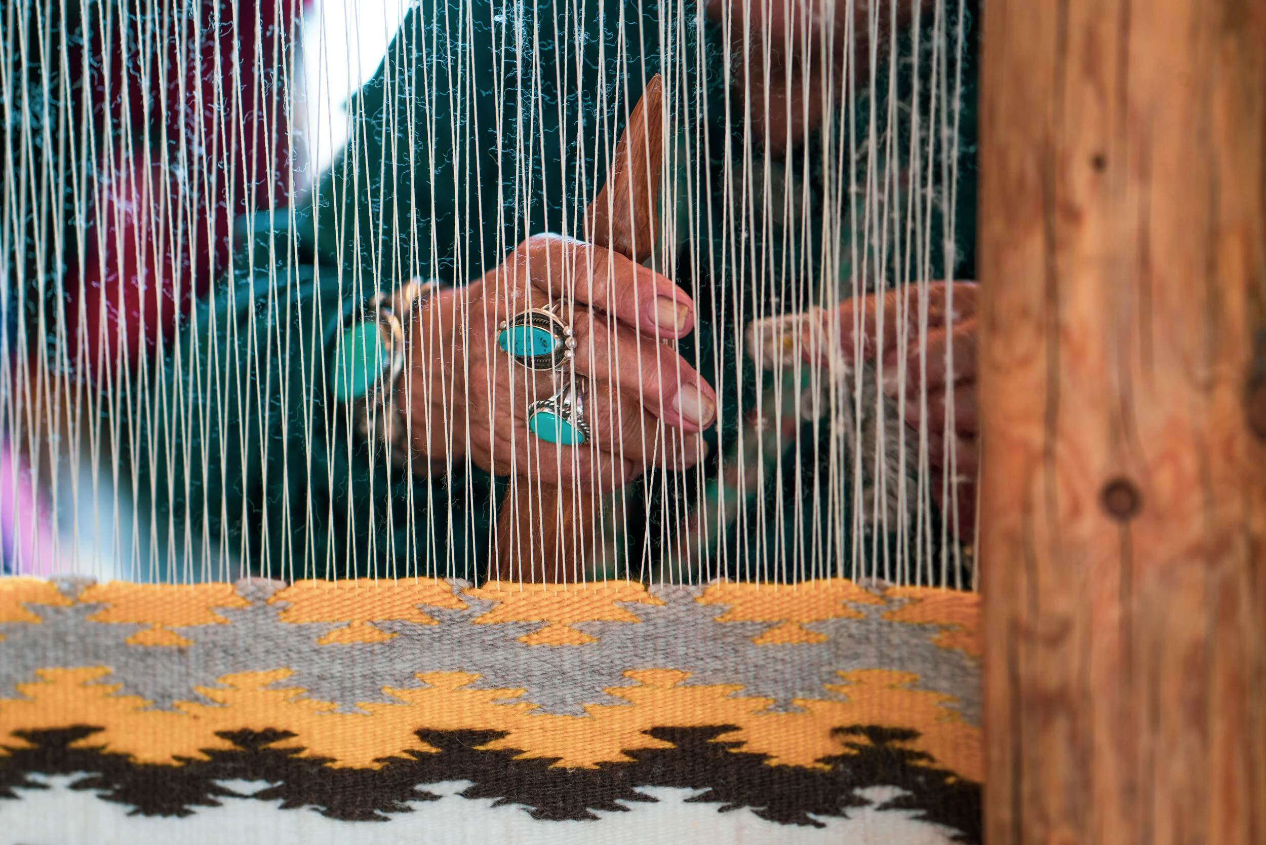 A person weaving on a loom, with various rings on their fingers, working with numerous vertical threads, and a colorful woven fabric at the bottom.