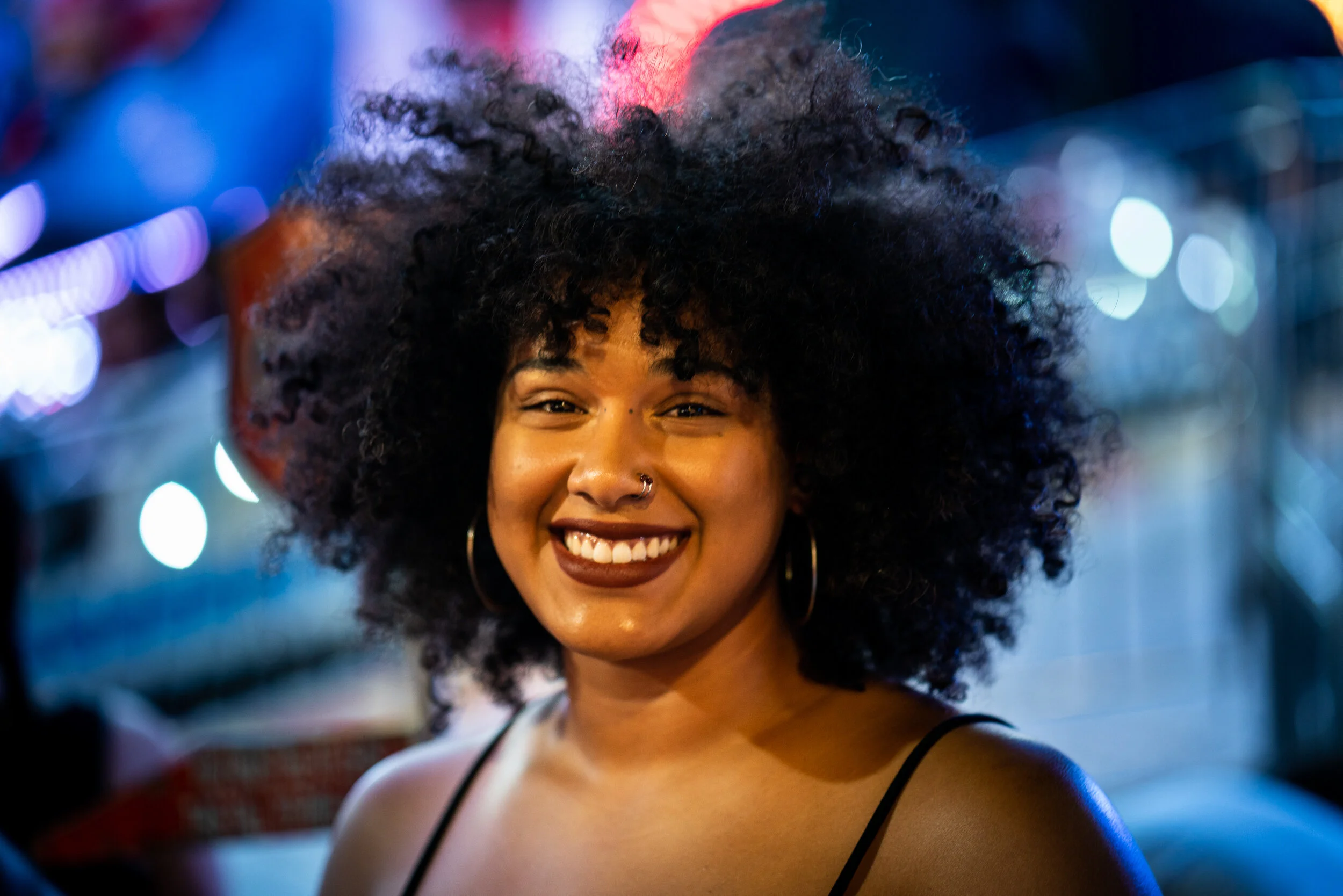 A smiling woman with curly hair, hoop earrings, wearing a black spaghetti strap top, in a vibrant, colorful nighttime setting with bokeh lights in the background.