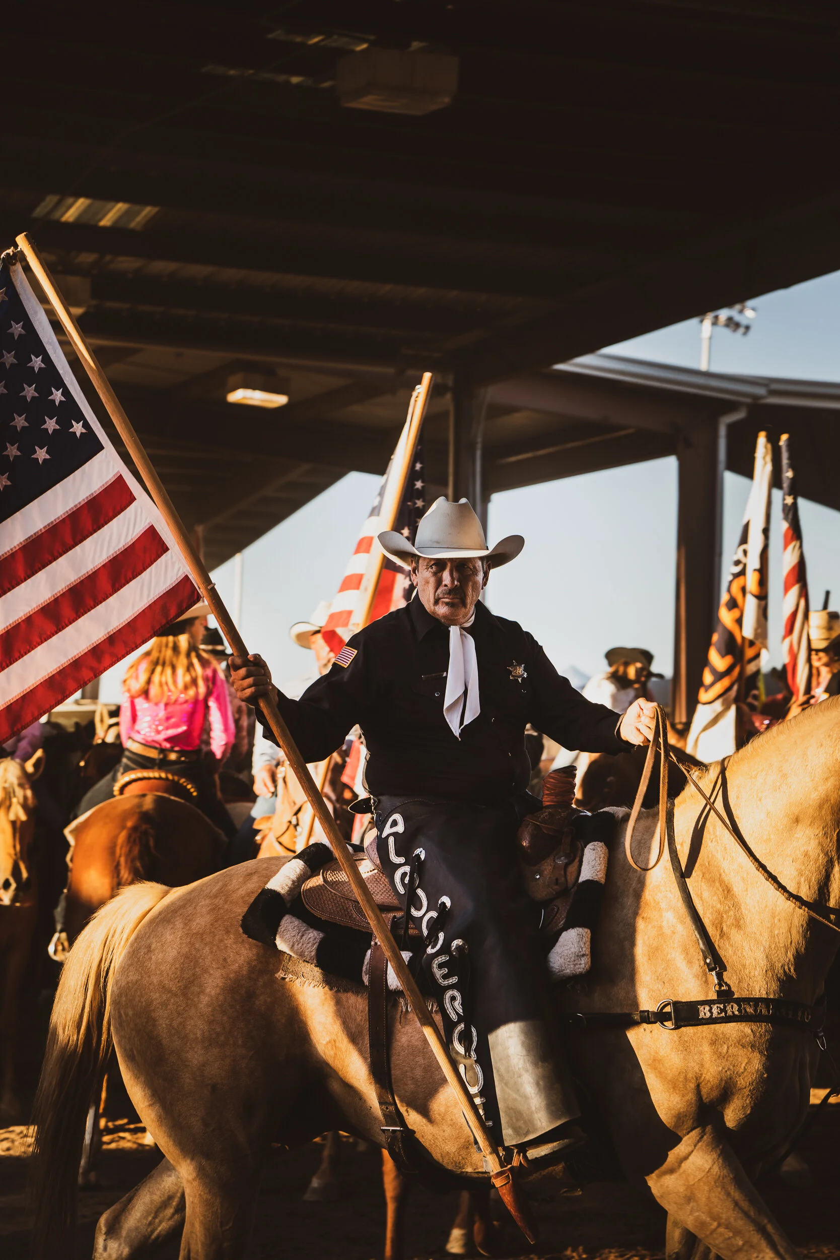 A cowboy riding a horse, holding an American flag, with other riders and flags in the background at a rodeo or parade.