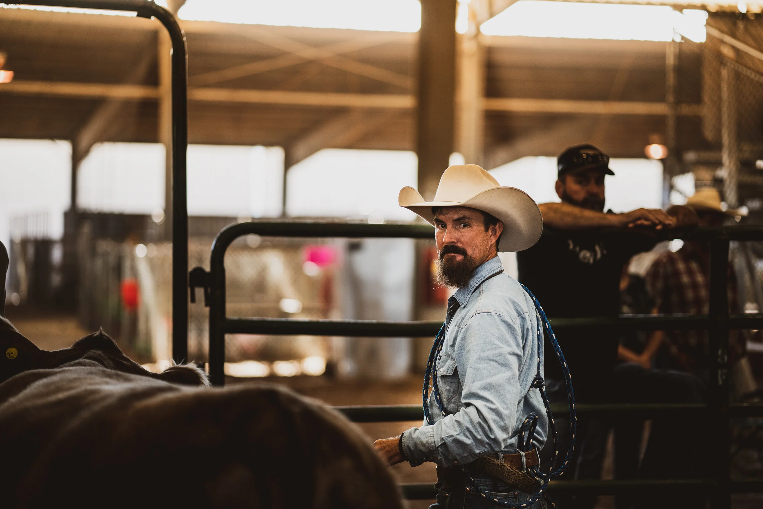 A man wearing a cowboy hat, jeans, and a denim shirt standing in a barn with other people in the background.