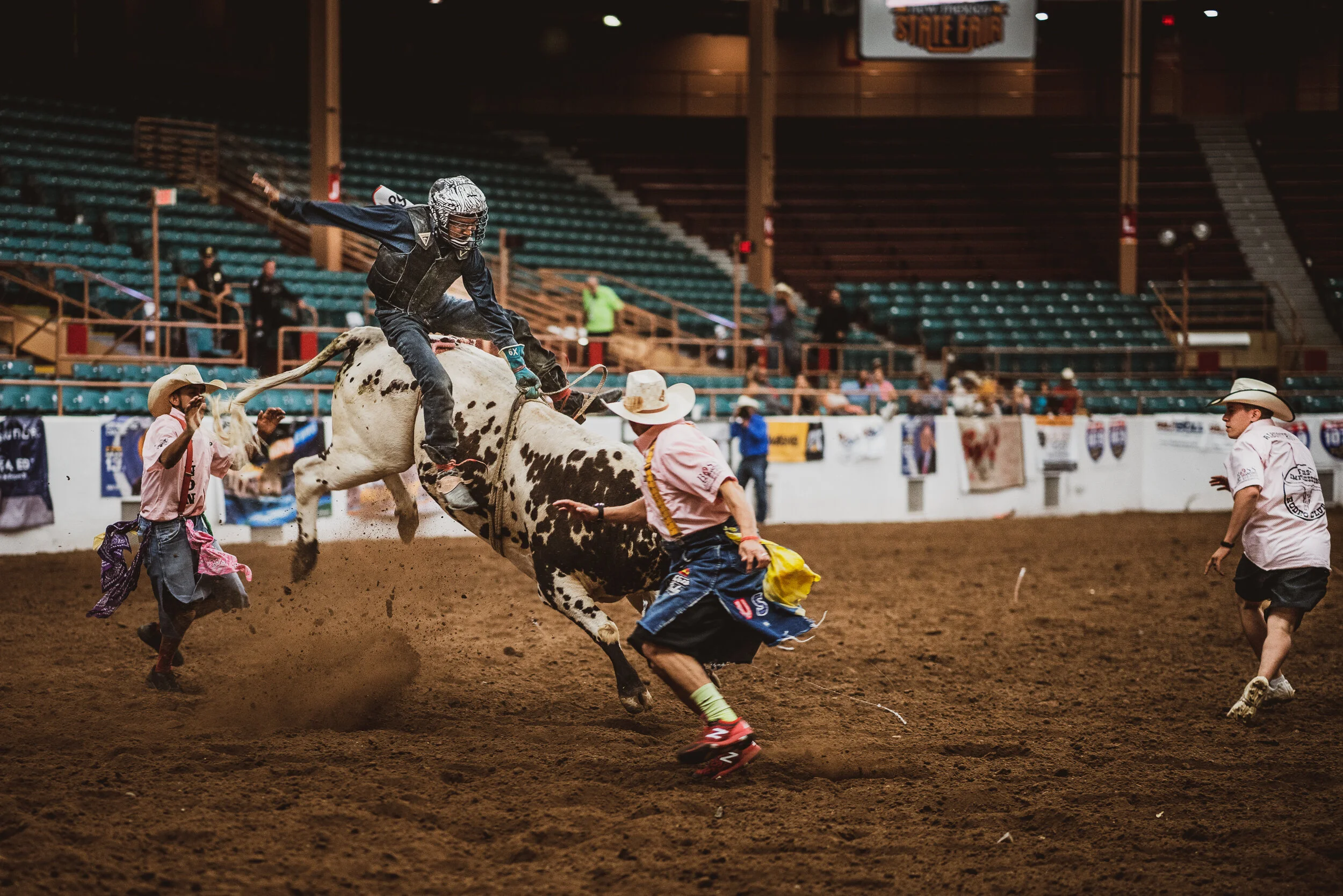 A rodeo event with a cowboy riding a bucking bull inside an arena. Several other rodeo participants in cowboy hats and attire are around the bull, with some running to help or control the animal. The arena has a dirt floor and empty bleachers in the 