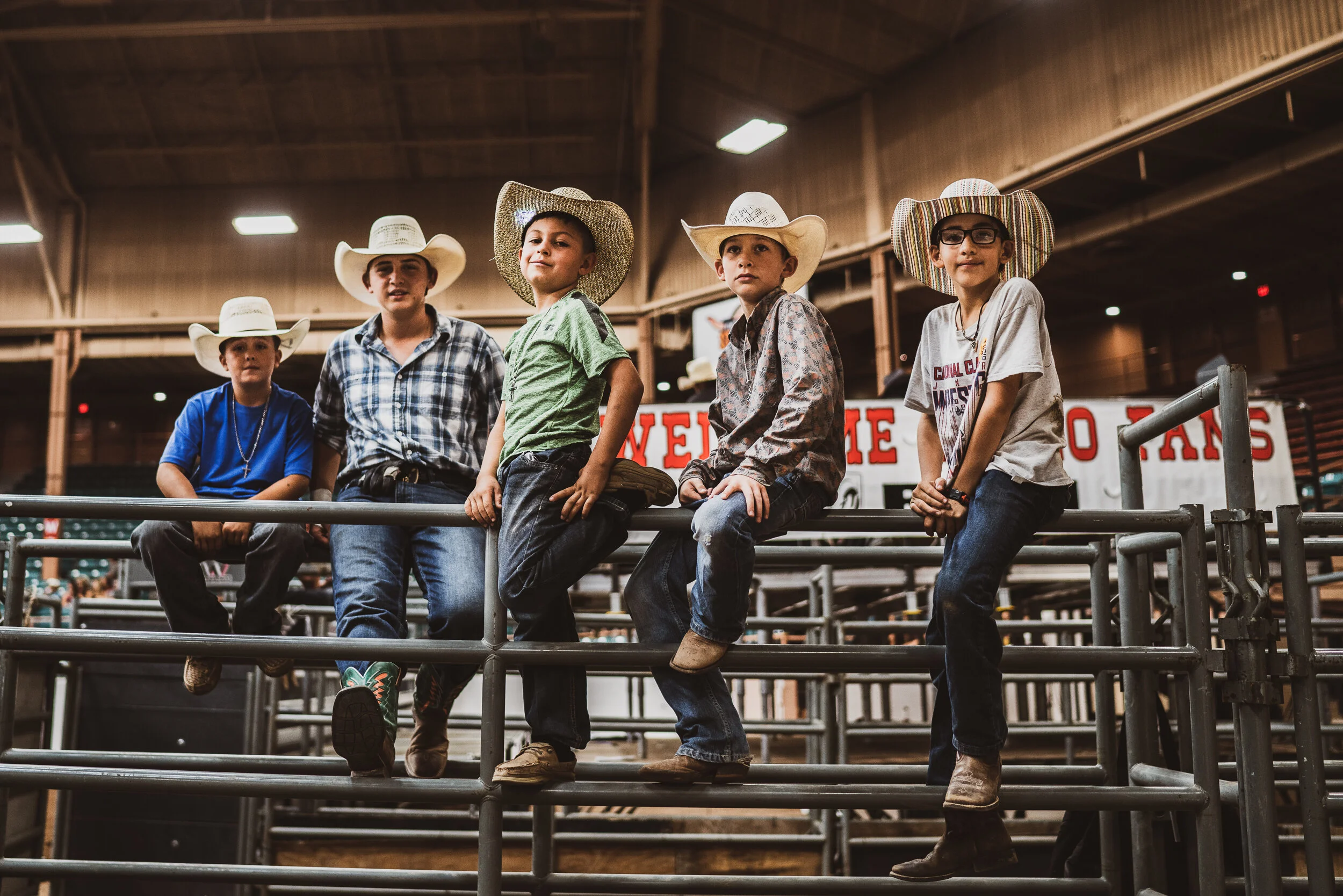 Five children in cowboy hats and casual clothes sitting on a metal railing inside a barn or arena, with a sign that reads 'Western Days' in the background. New Mexico State Fair