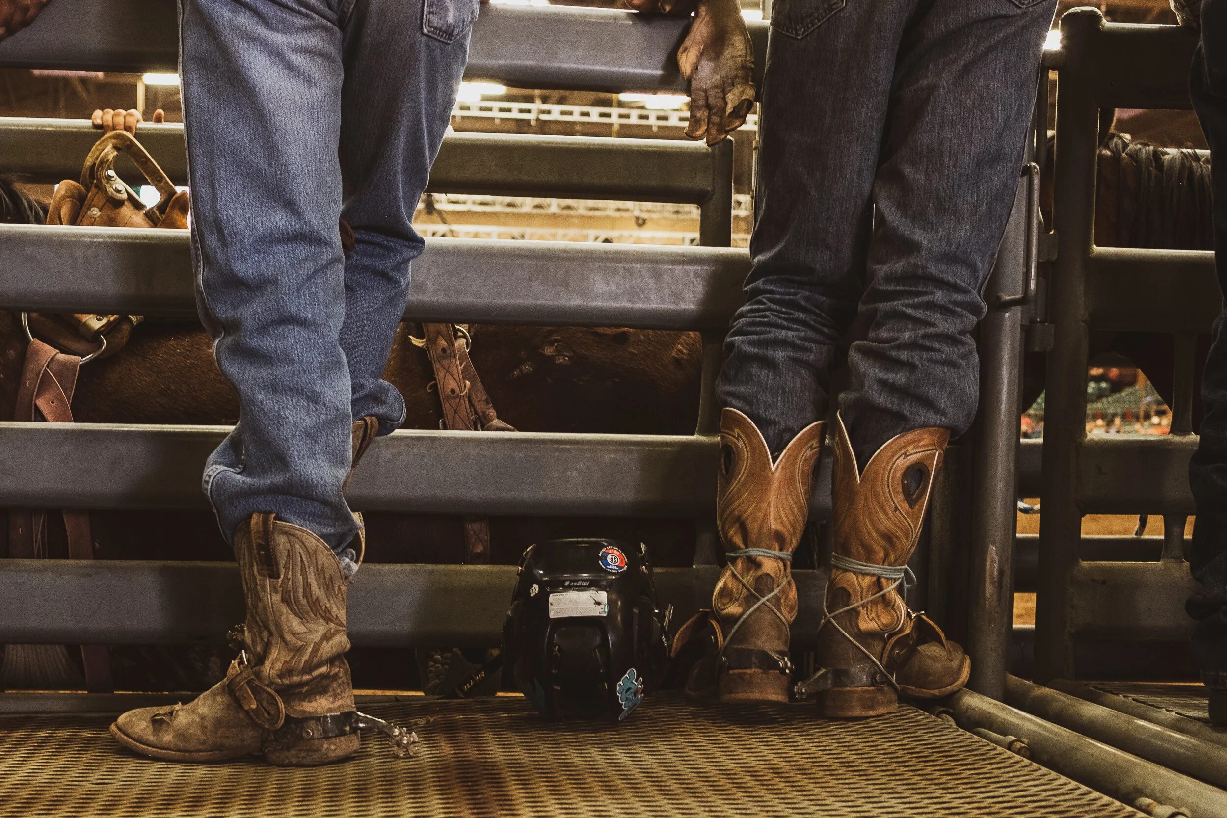 View of two people wearing jeans and cowboy boots standing on a metal platform inside a rodeo arena, with a helmet and horse tack in the background. New Mexico State Fair
