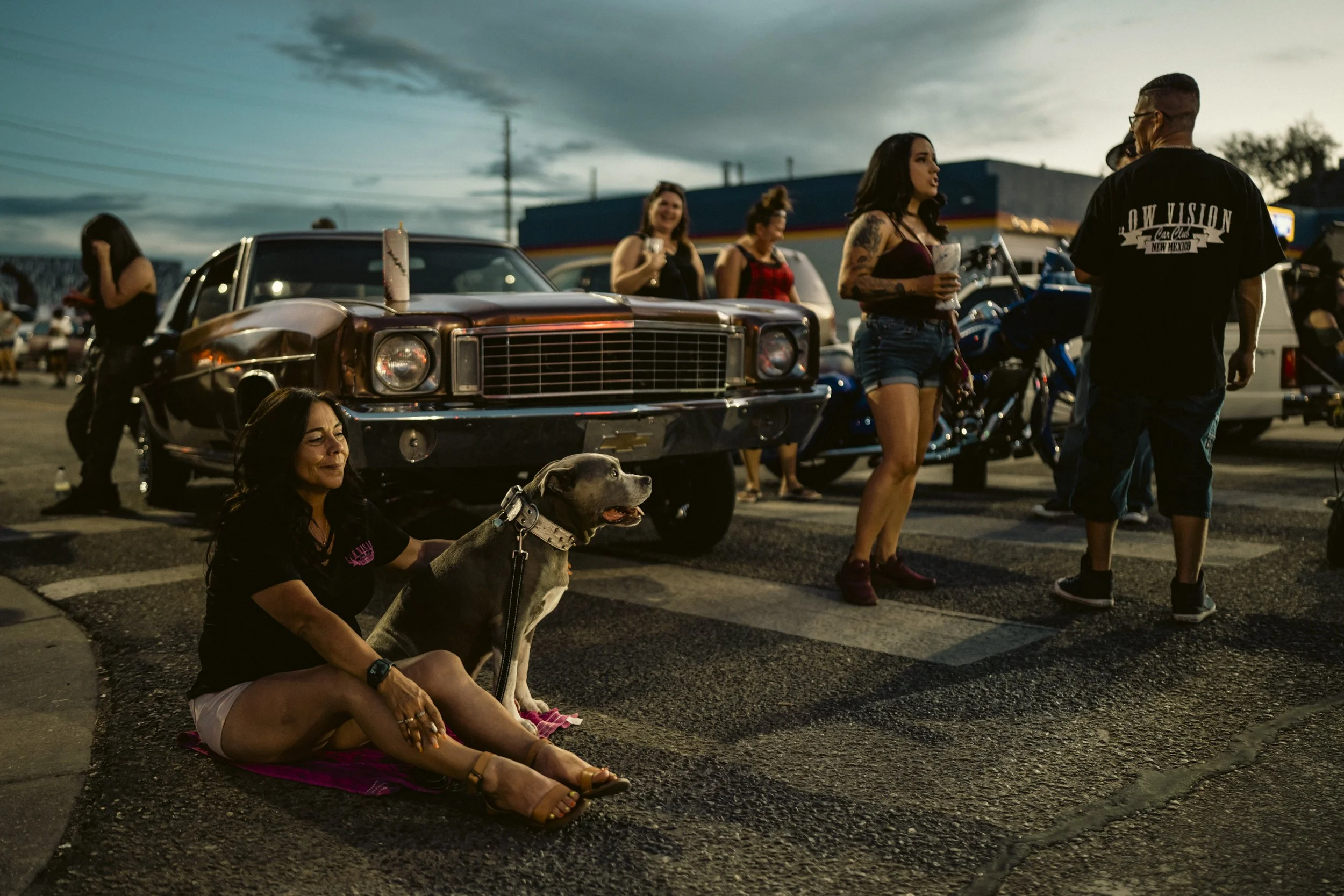 People gather around a classic lowrider at dusk in downtown Albuquerque while a woman sits with a dog in the foreground.
