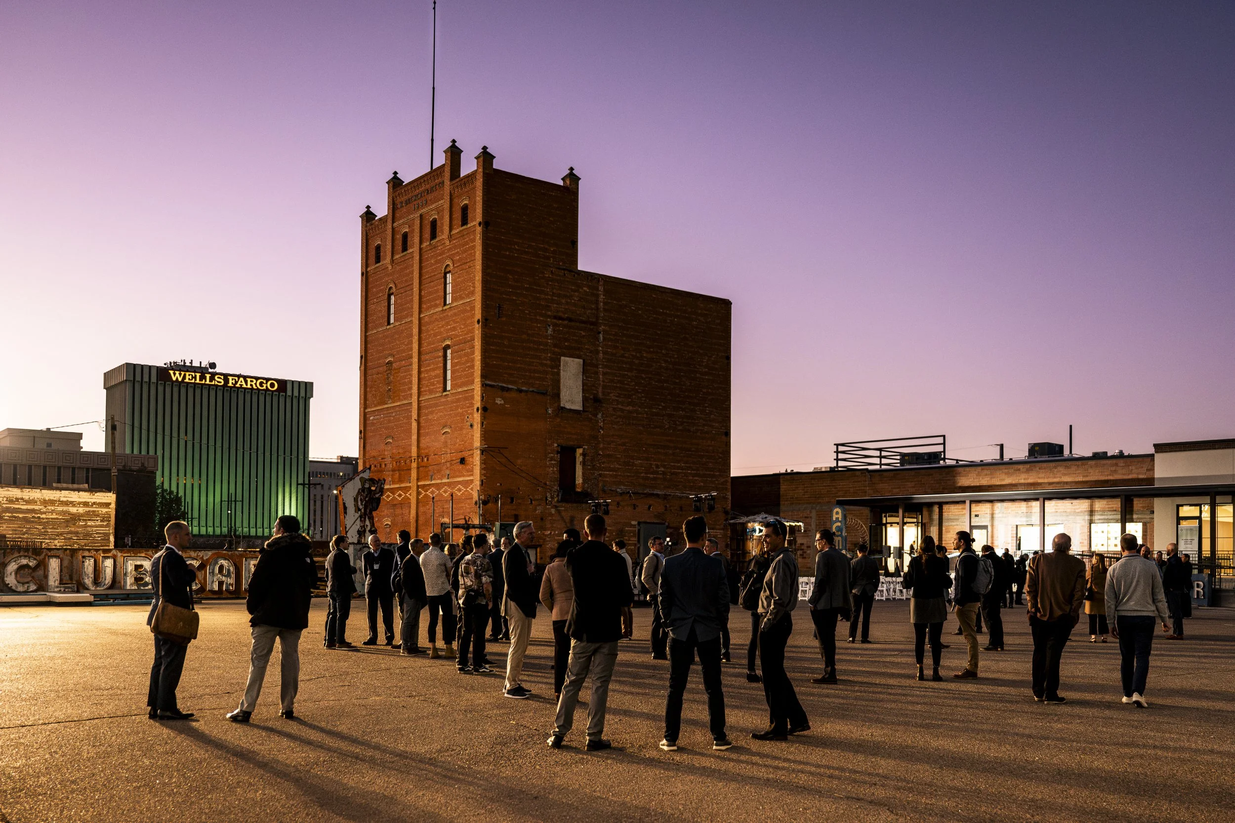 Group of people standing outdoors in front of a brick tower and modern buildings during sunset.